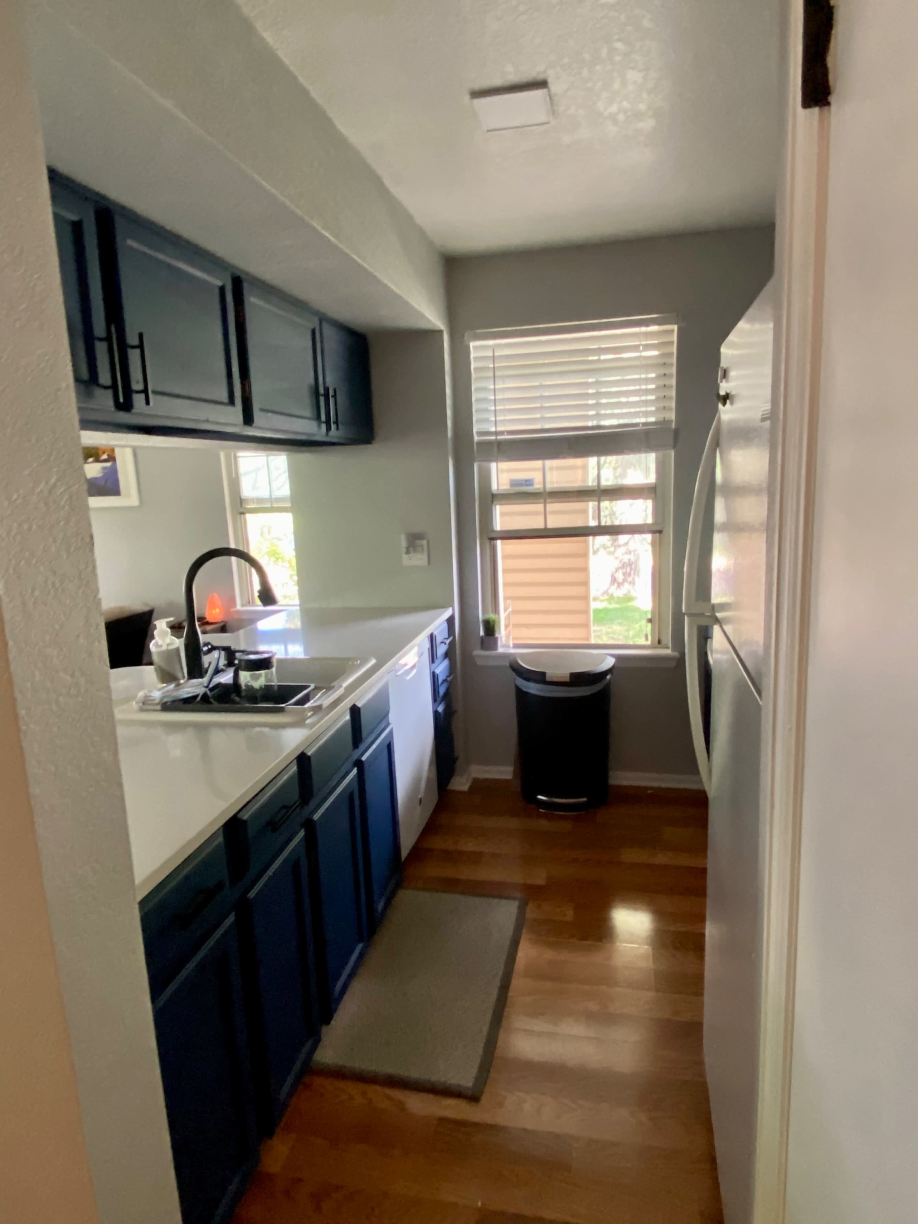 A narrow kitchen features blue cabinets, a white countertop, a window beside the sink, and a trash can near the refrigerator.