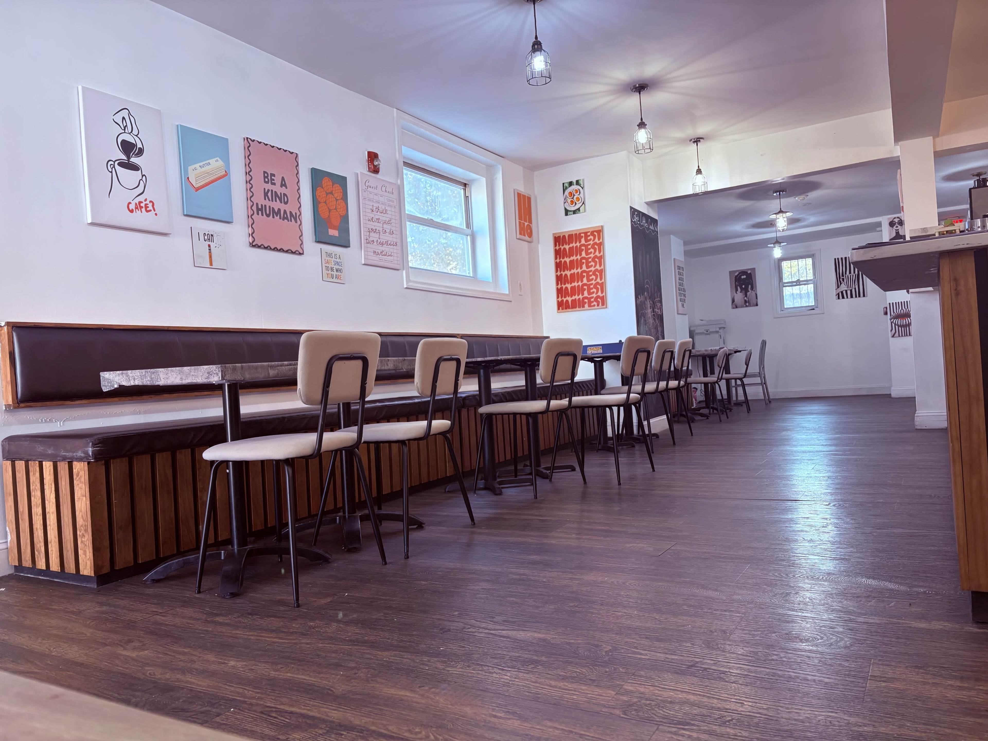 The image shows a modern cafe interior with wooden flooring, several empty tables and chairs, and colorful wall art.