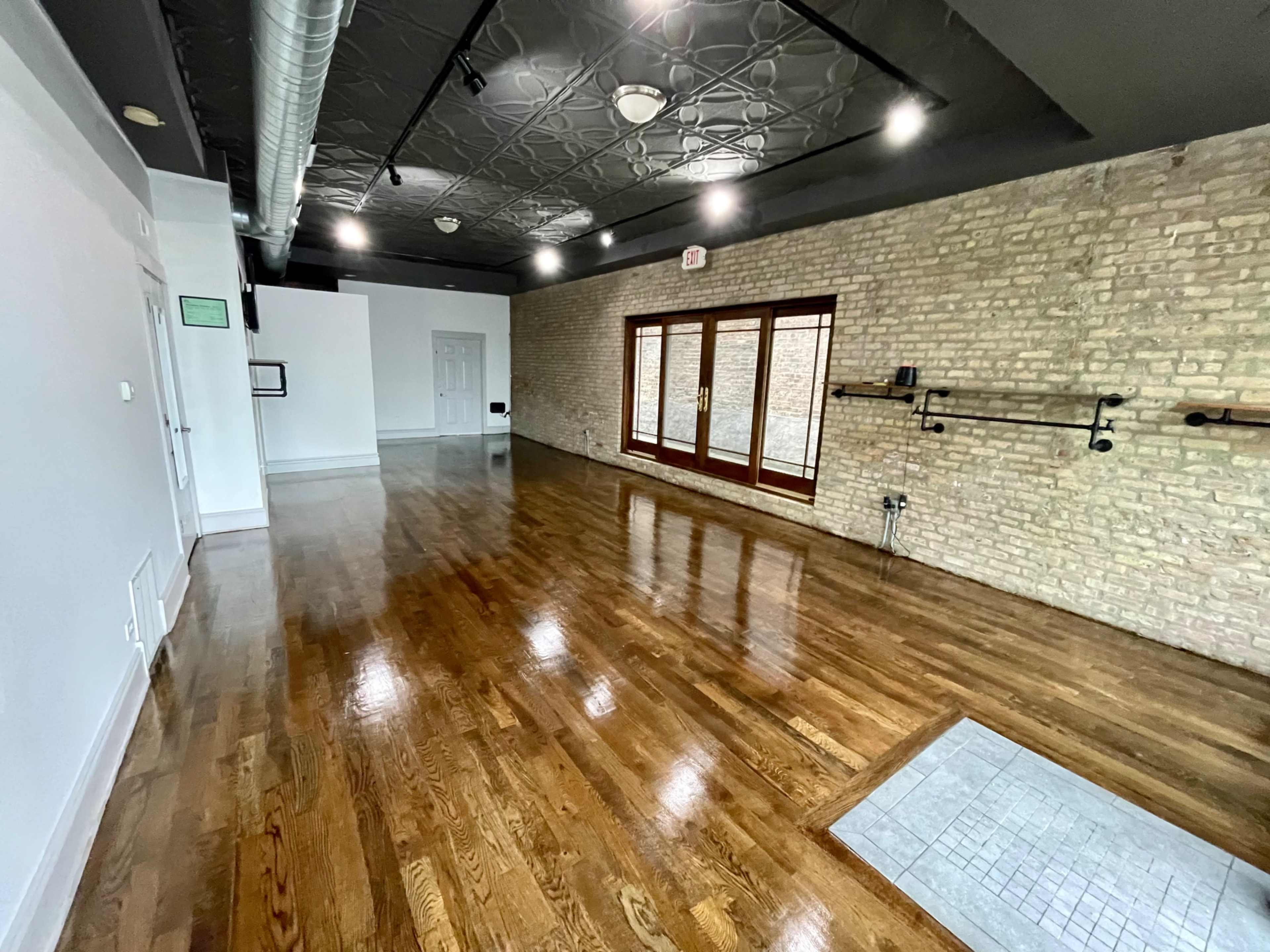 An empty room with hardwood flooring, exposed brick walls, and large windows along one side.