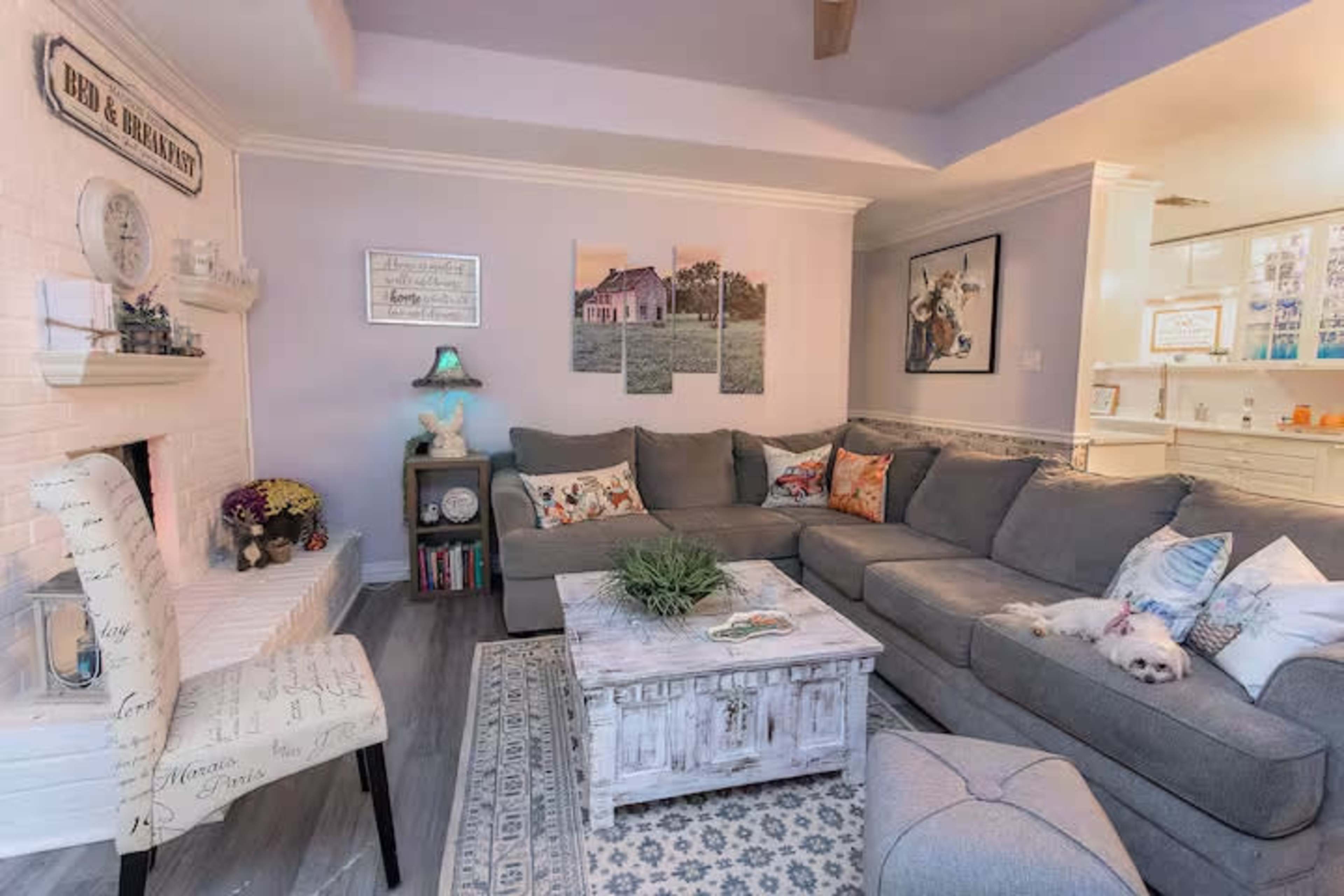 The image shows a cozy living room featuring a large gray sectional sofa, a rustic coffee table, and a white fireplace accented with decorative items.