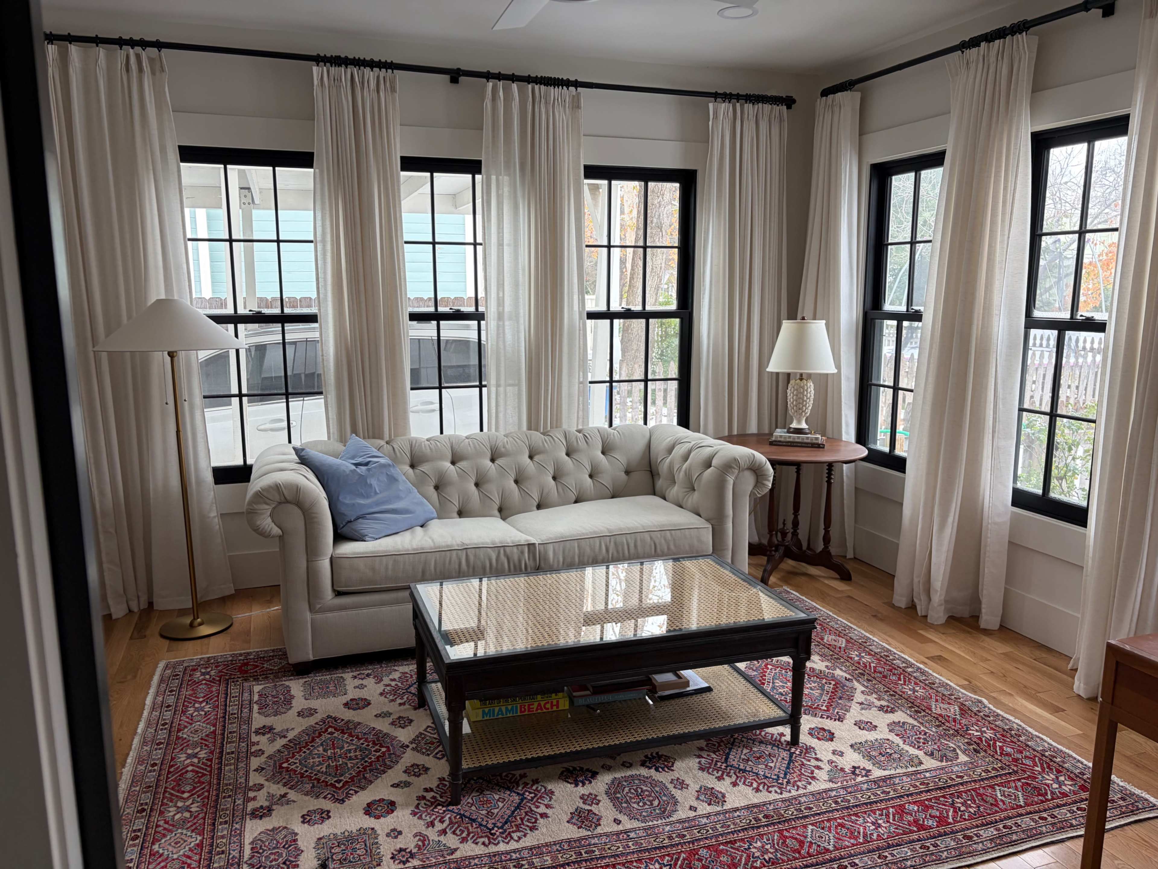 The image shows a bright, cozy living room featuring a white tufted sofa, a wooden side table with a lamp, and a patterned rug on the hardwood floor.