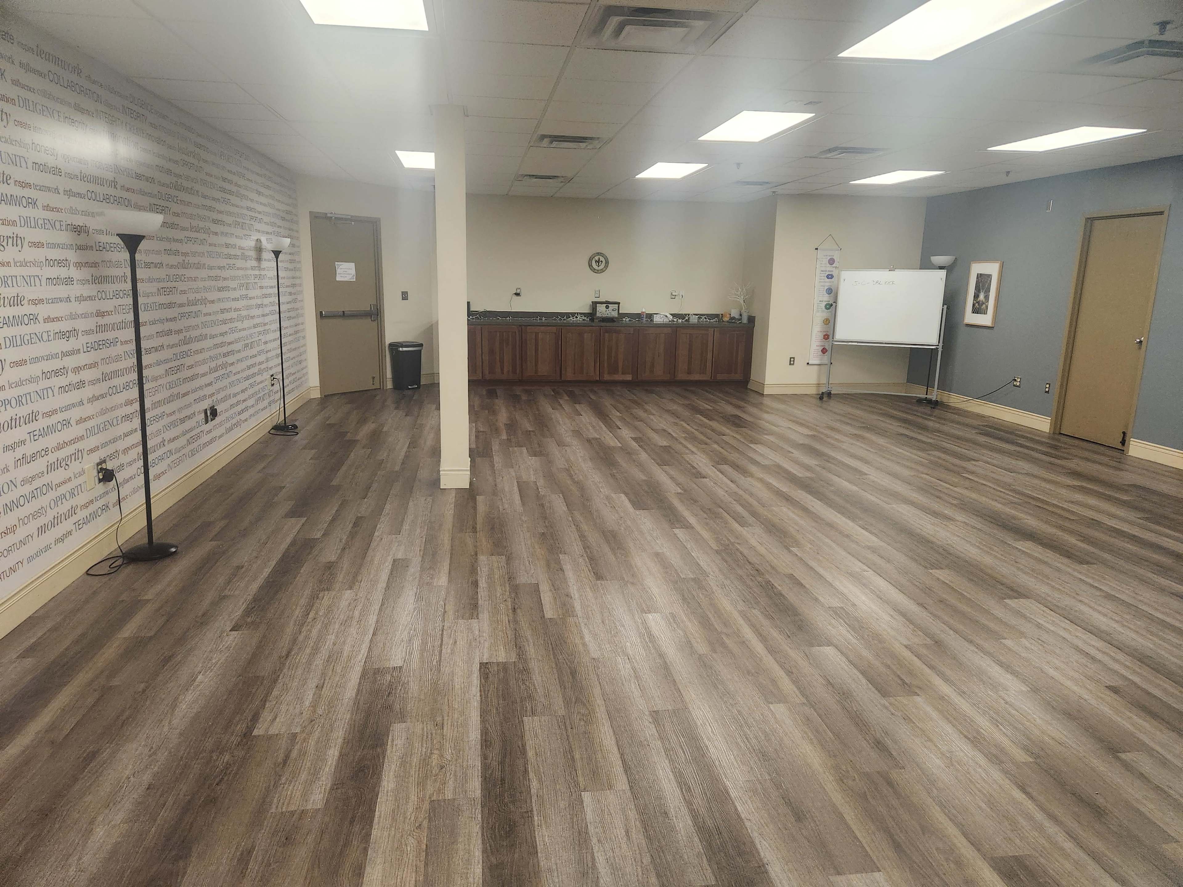 The image shows a spacious room with wooden flooring, a kitchenette area in the background, and a whiteboard on wheels along one side.