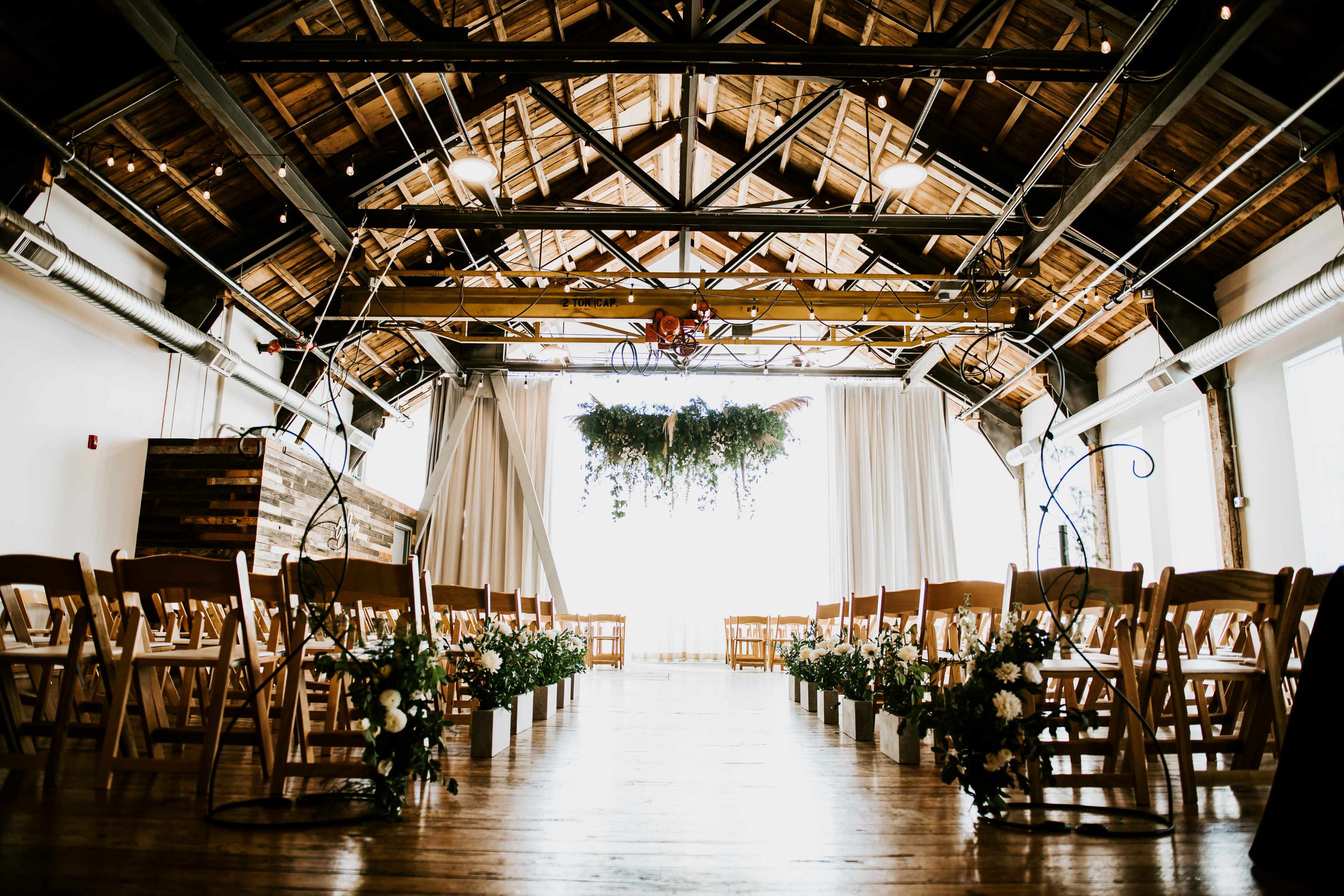 A spacious venue with wooden beams features rows of chairs set for a wedding ceremony, adorned with floral arrangements and hanging greenery.