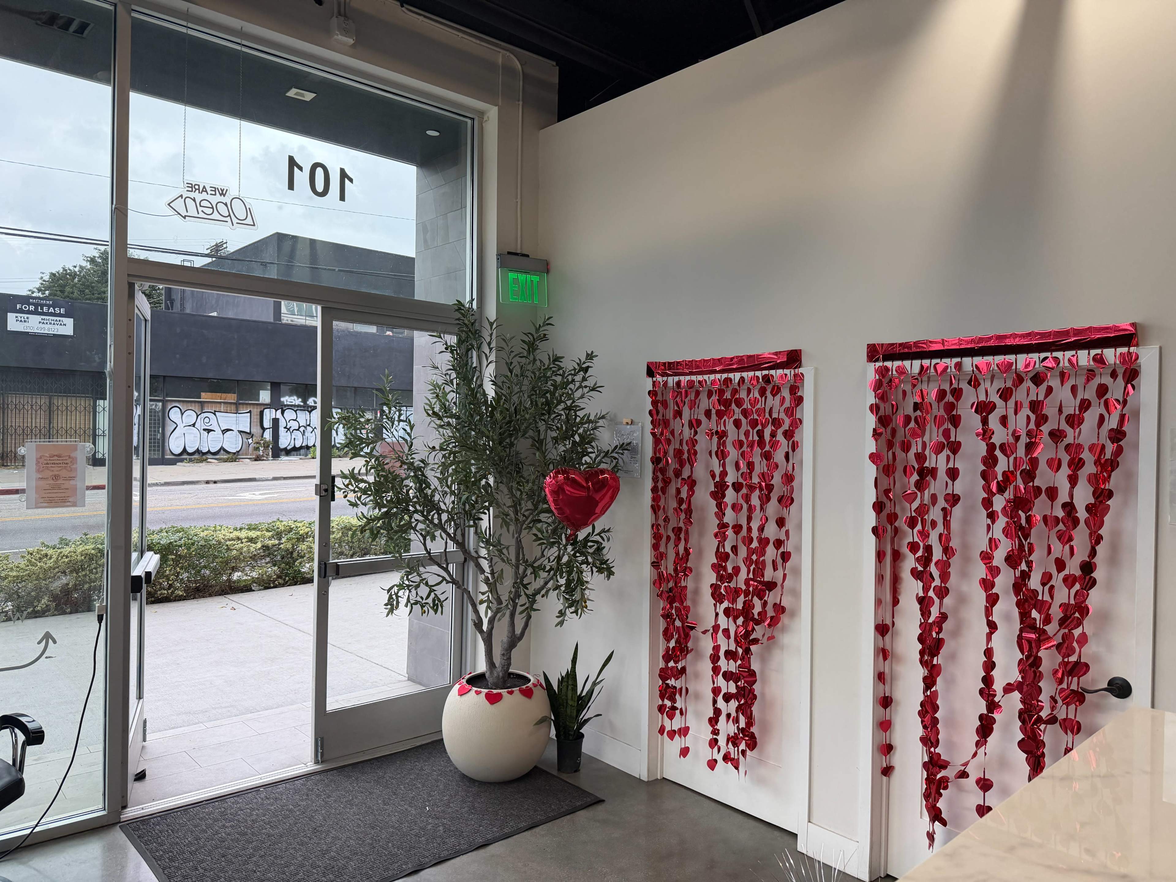 The interior of a shop features a large entrance with an open door, a decorative wall adorned with red hearts and strings, and a potted tree in the corner.