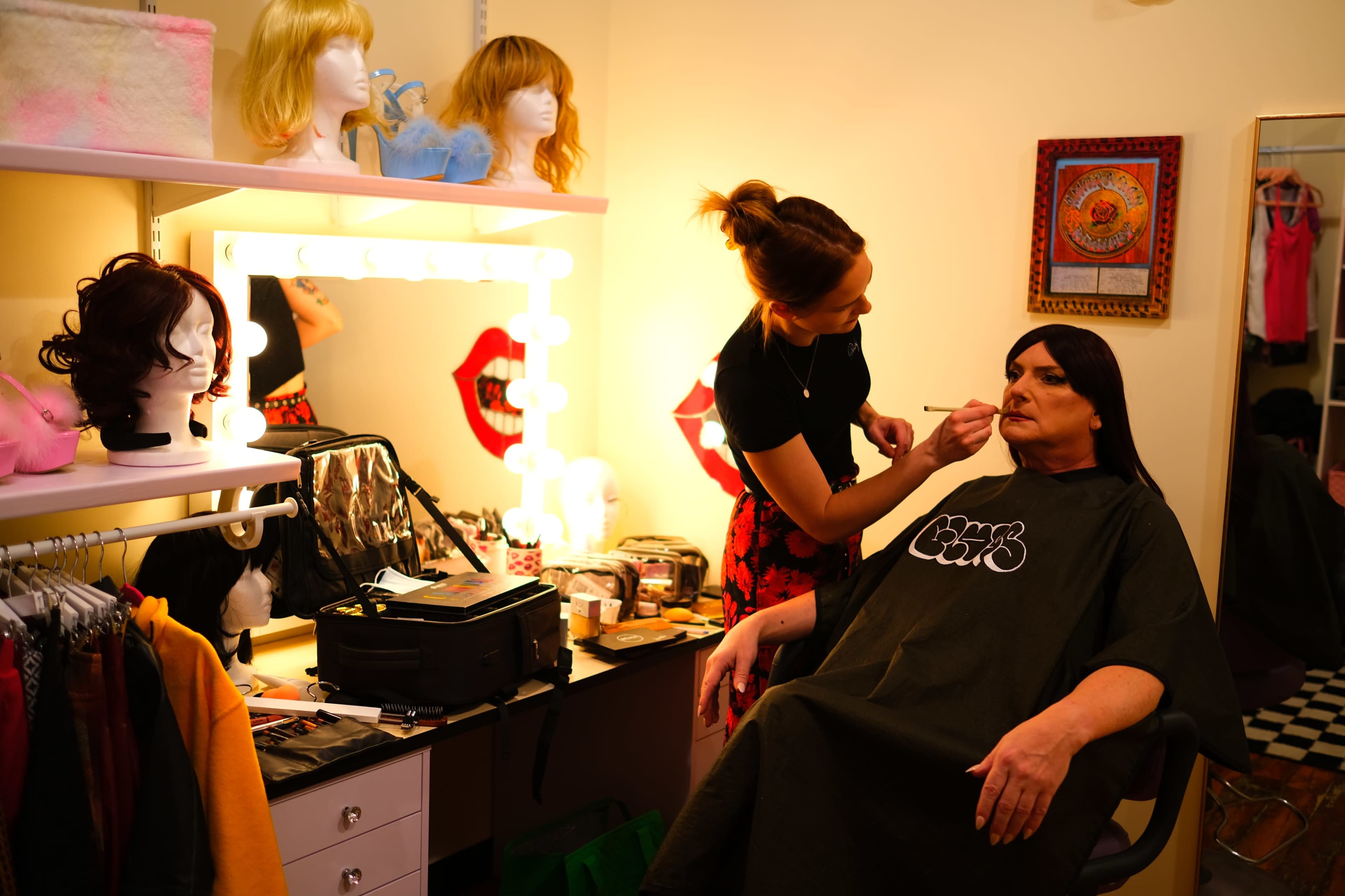 A person sits in a chair receiving makeup application in a room decorated with mannequin heads and a brightly lit vanity.