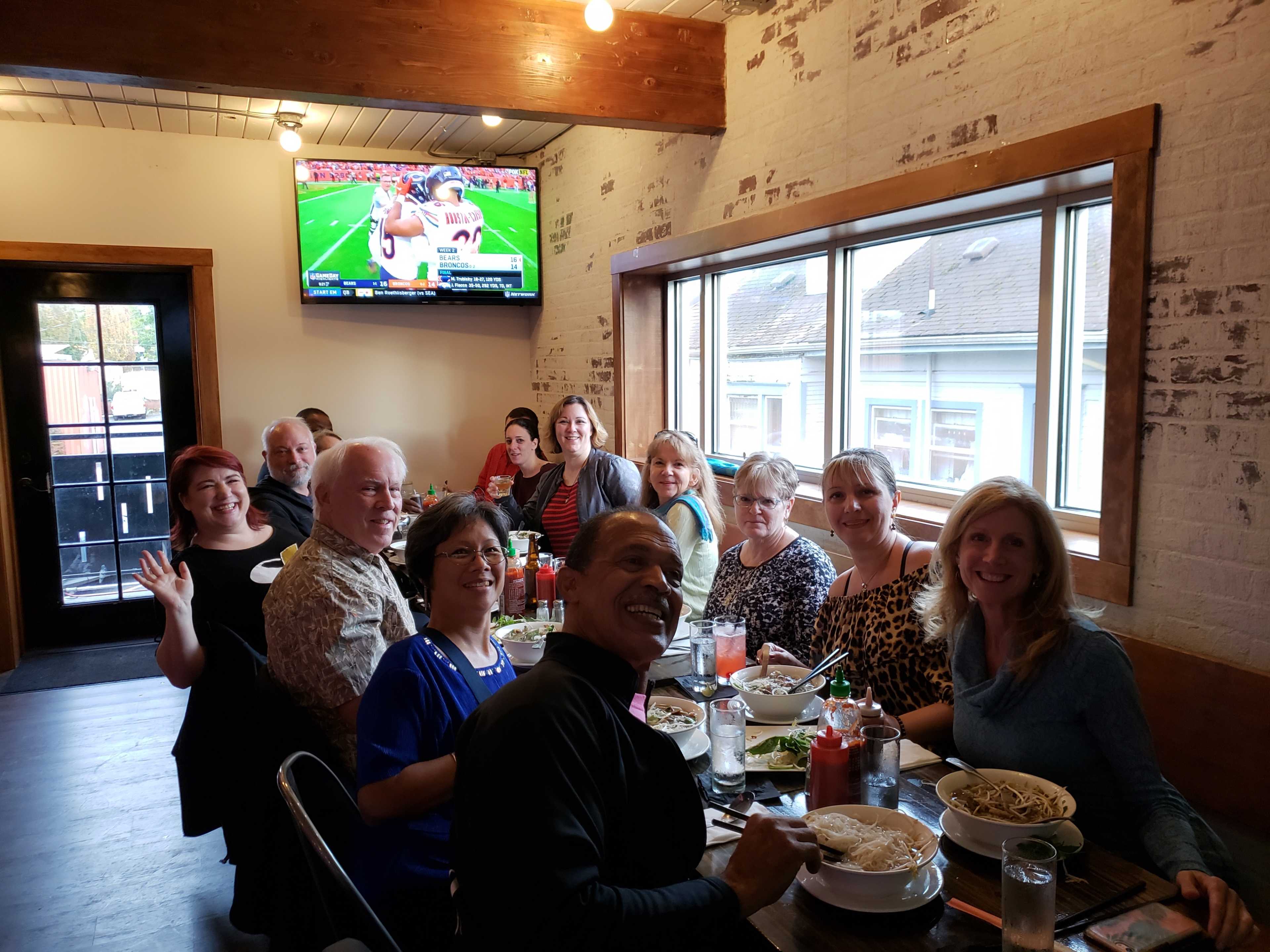 A group of twelve people is gathered around a dining table in a restaurant, with a television displaying a football game in the background.