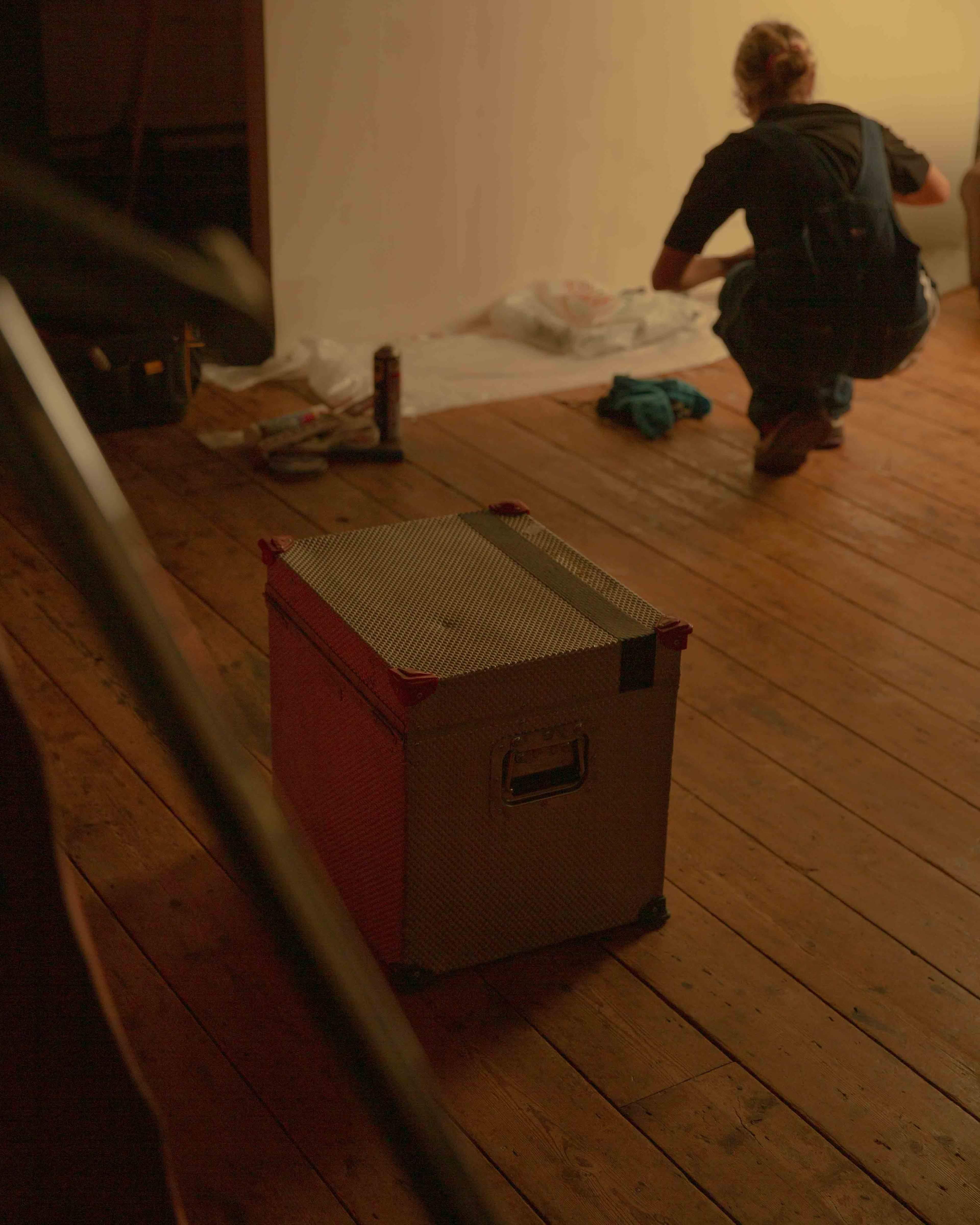 A metal storage box sits on a wooden floor while a person works in the background.