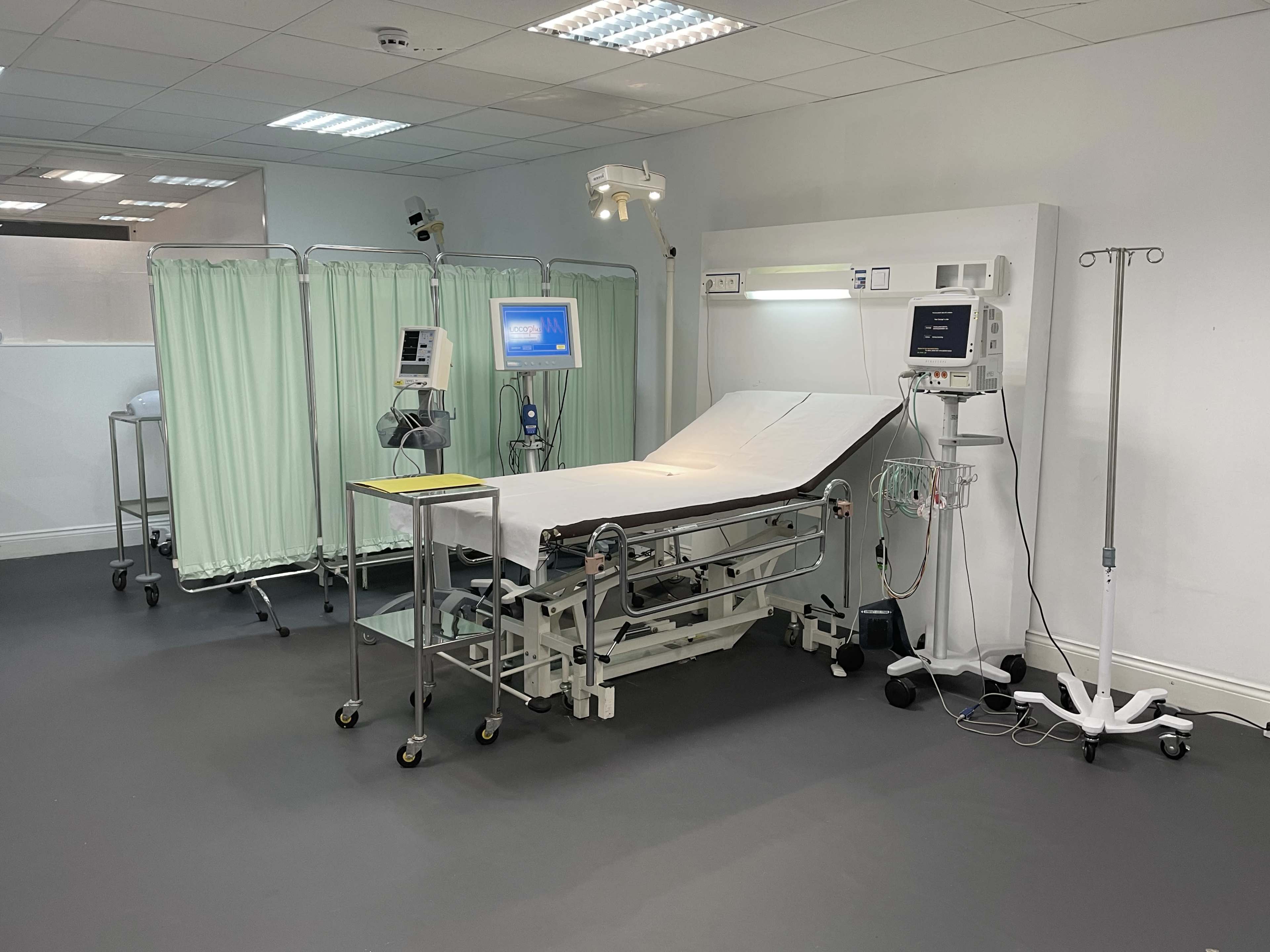 A medical examination room with an adjustable examination table, medical monitors, and green privacy curtains.