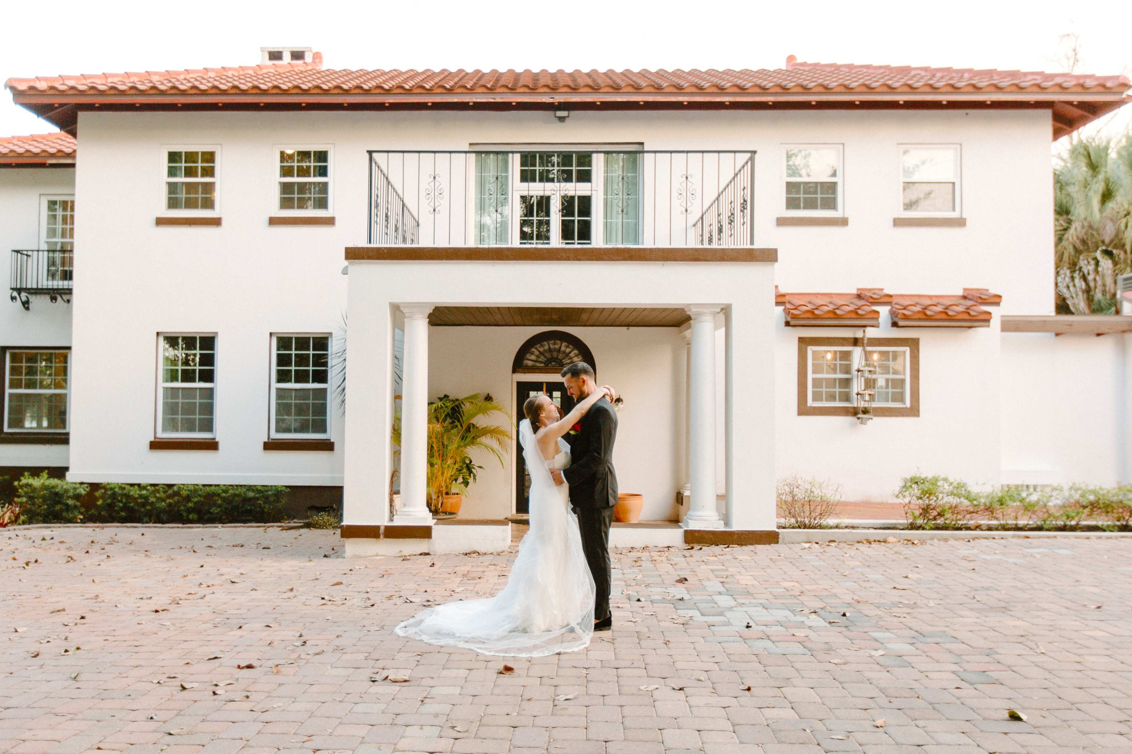 A bride and groom embrace in front of a large white house with a red-tiled roof and a stone pathway.