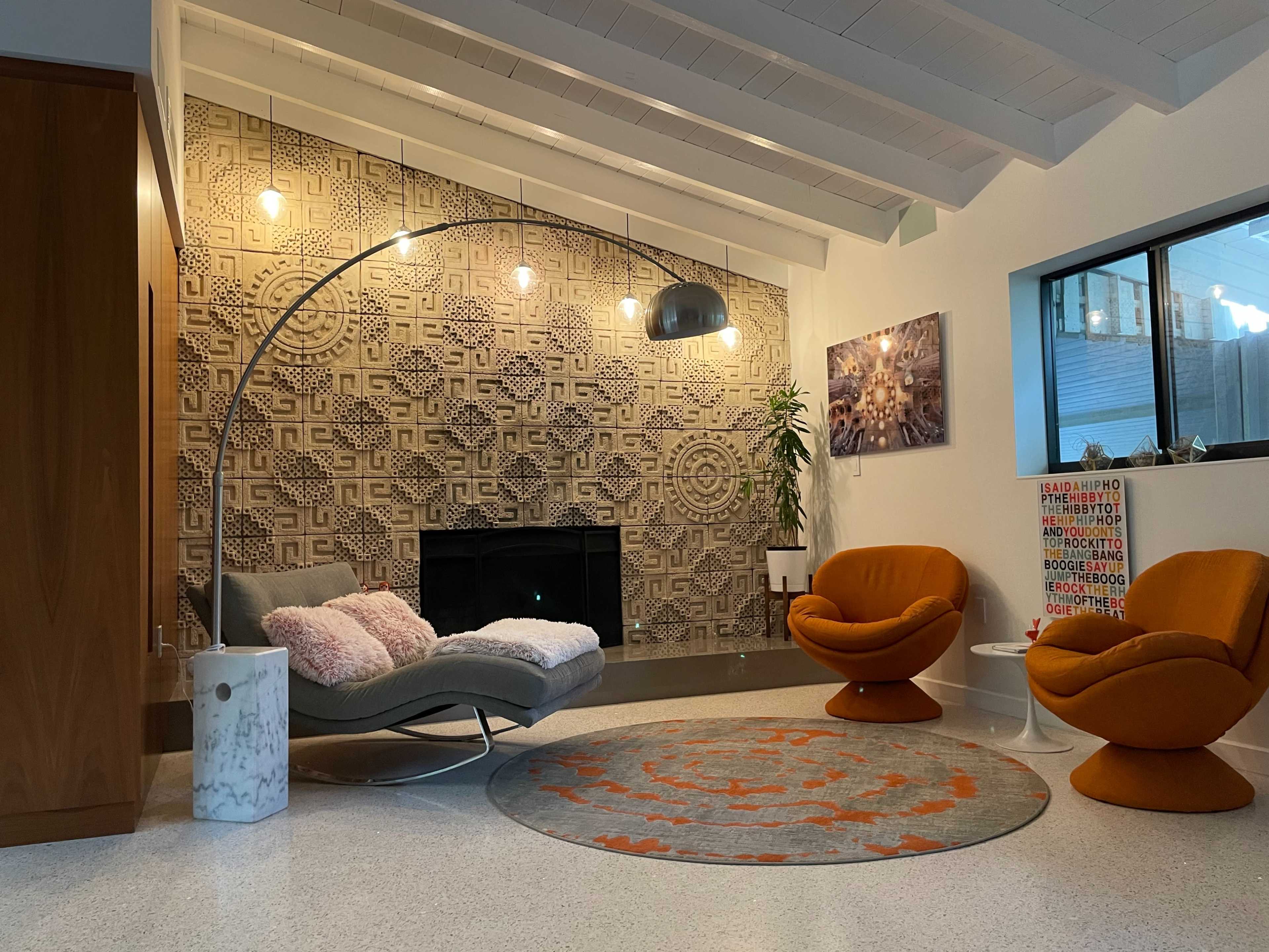 A modern living room featuring two orange lounge chairs, a curved gray chaise, a patterned wall, and a round area rug on a light-colored floor.