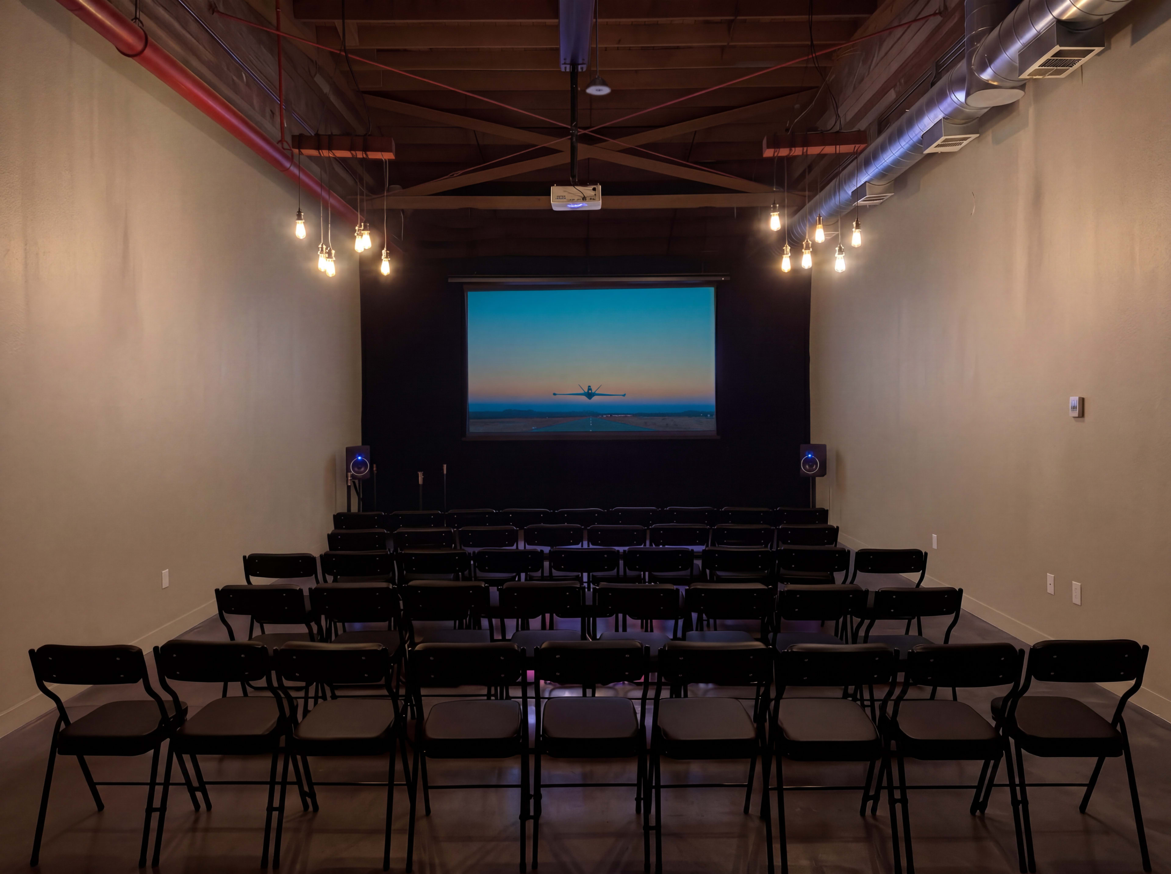The image shows a small, empty theater with rows of black chairs facing a screen displaying an image of an airplane in the sky.