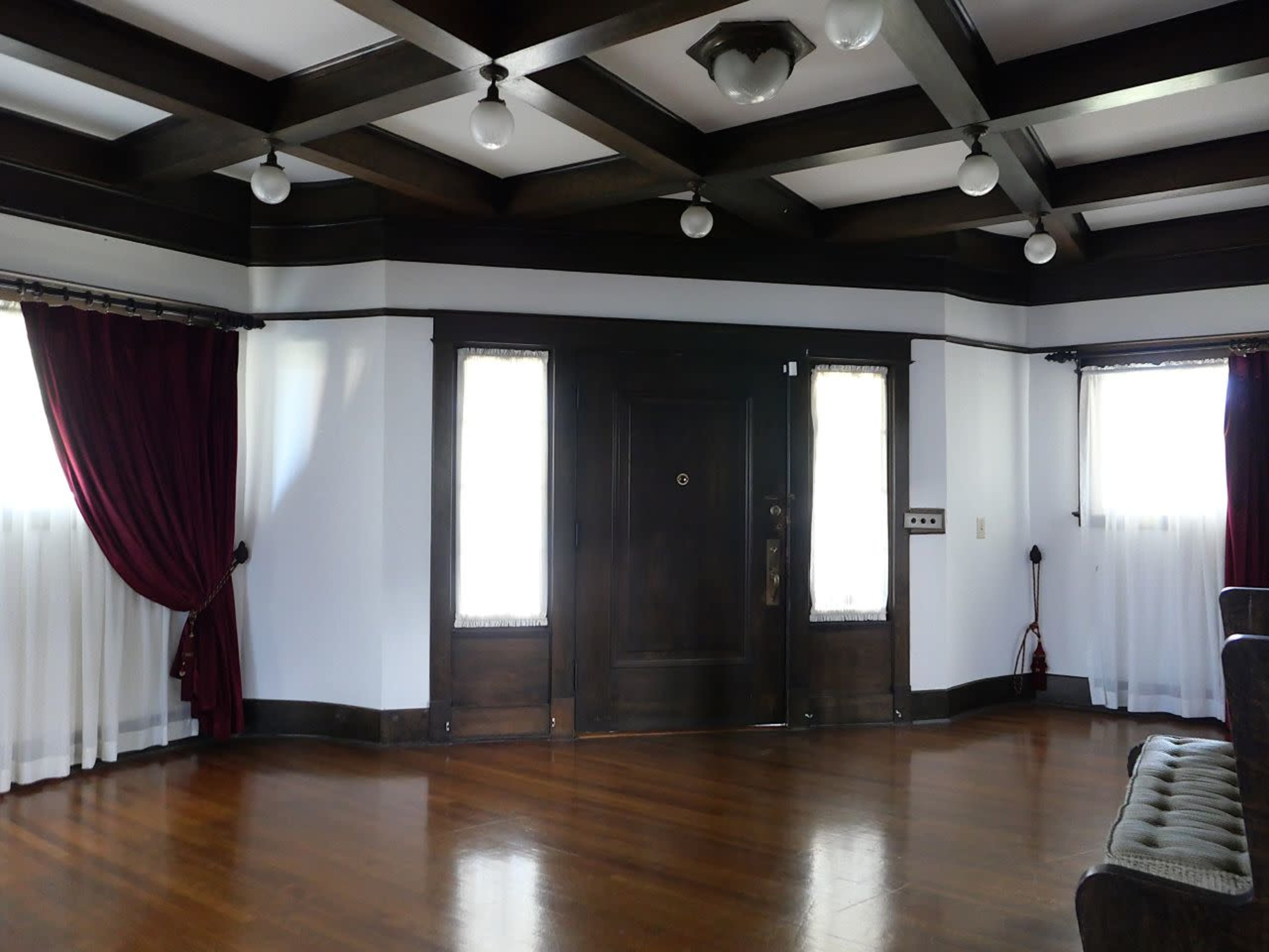The image shows a room with hardwood floors, a dark wood ceiling with beams, and windows dressed in white curtains and red drapes.