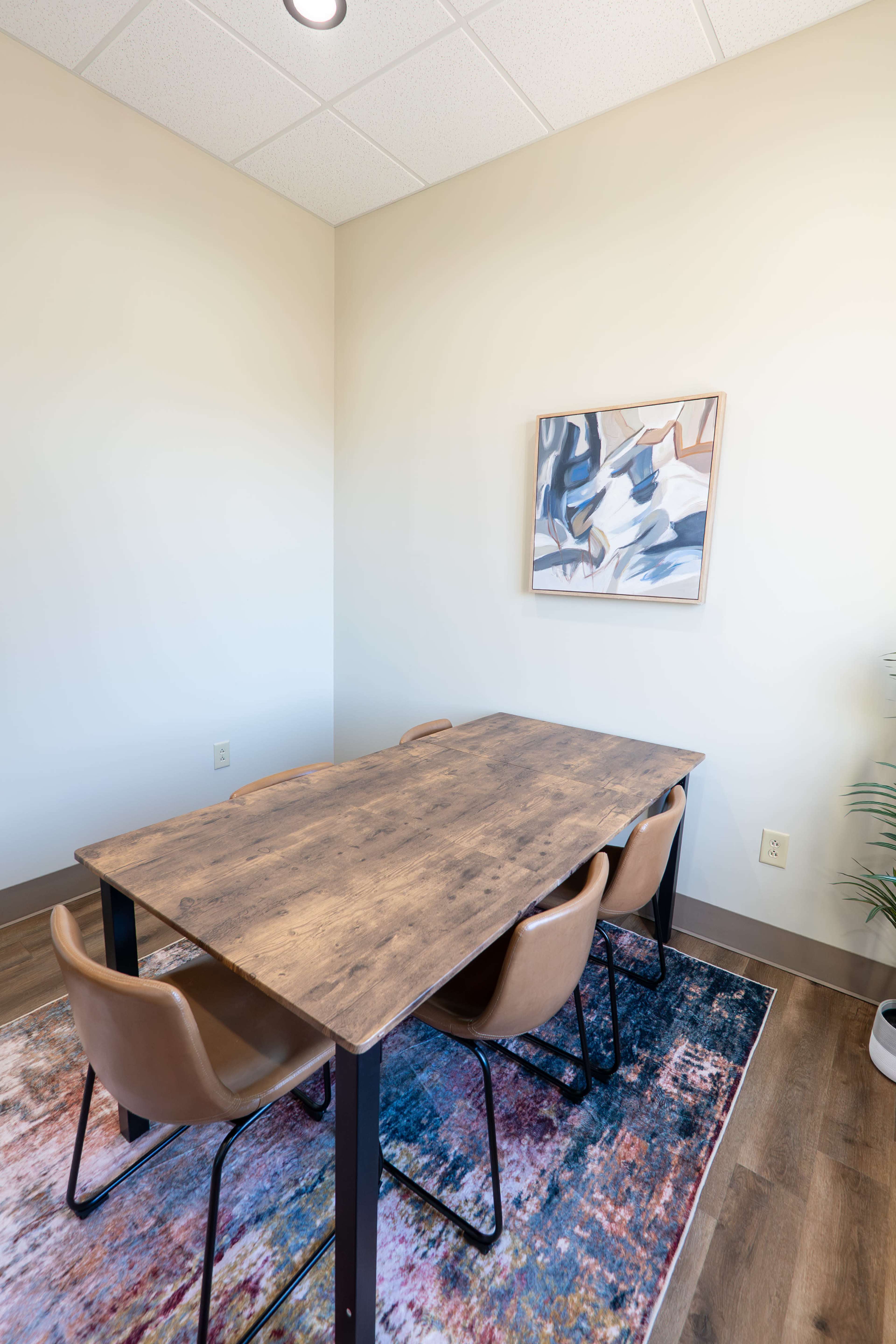 A meeting room features a wooden table surrounded by four brown chairs, with a colorful rug and abstract artwork on the wall.