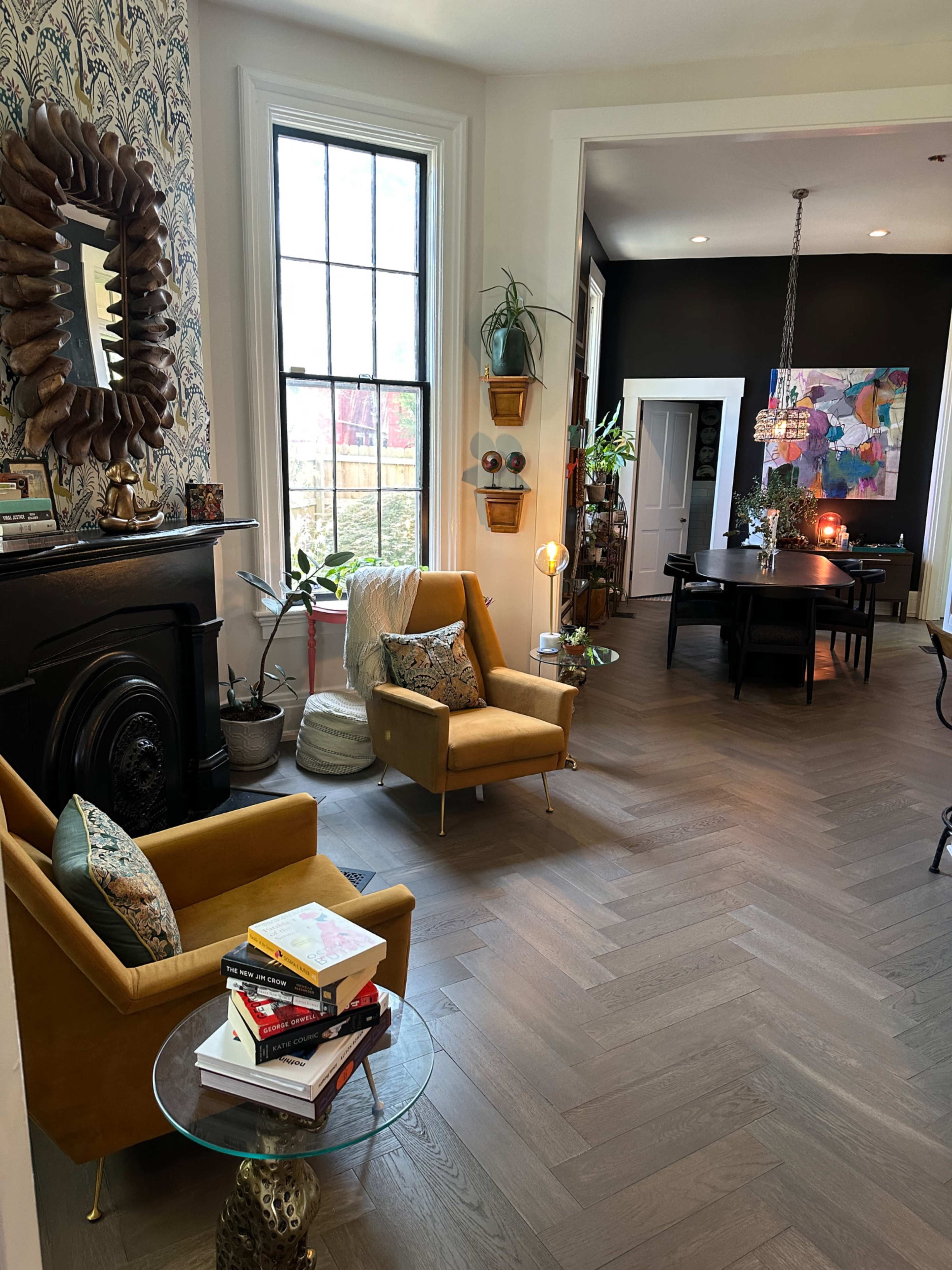 The image features a cozy living space with two yellow armchairs, a glass side table stacked with books, and a dining area in the background.