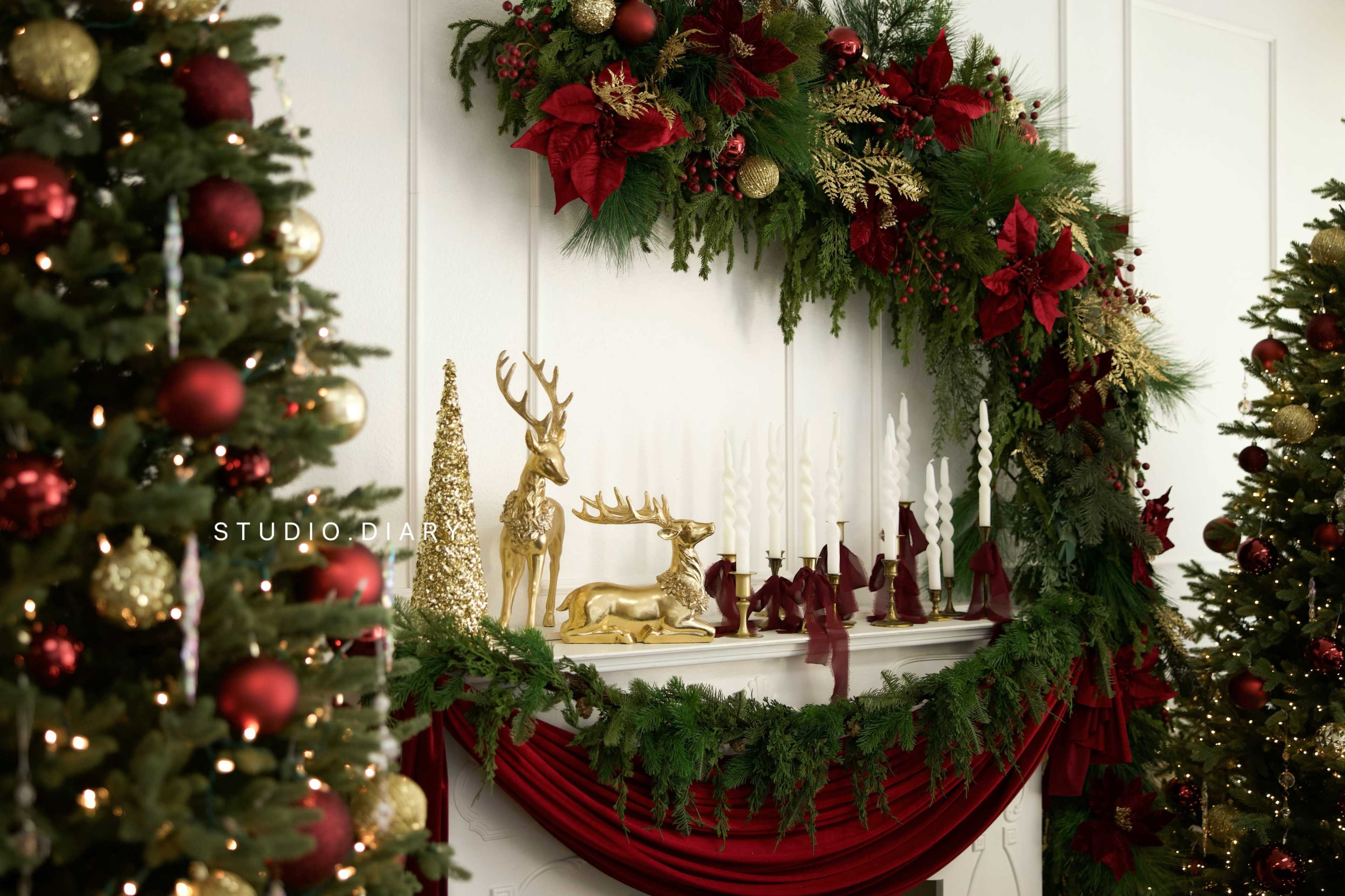 The scene features a festive mantel decorated with lush greenery, red floral accents, golden reindeer figures, and candlesticks, framed by Christmas trees adorned with ornaments.