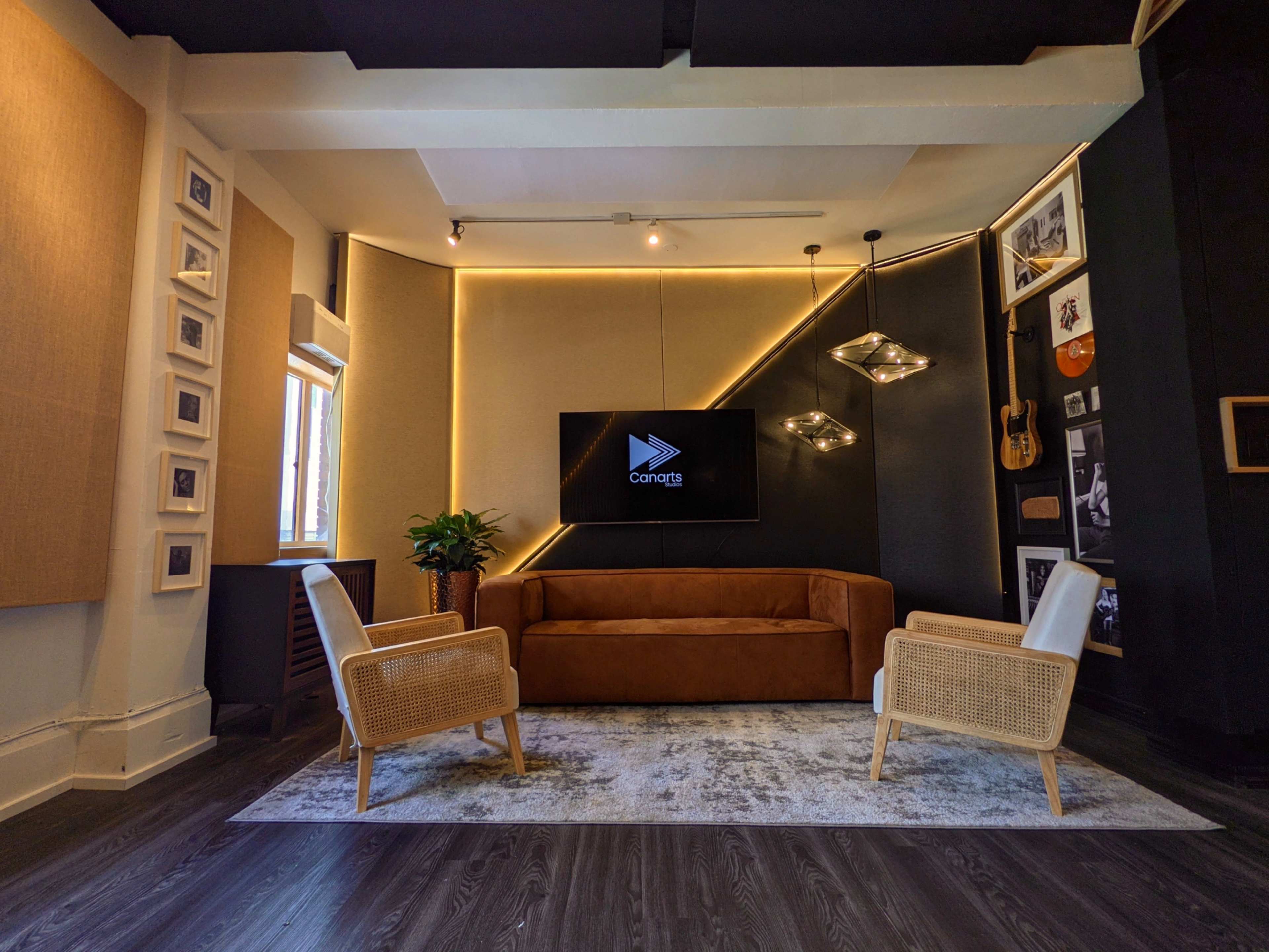 A modern living room with a brown sofa, two light-colored chairs, a television on the wall, and decorative elements including a plant and framed pictures.