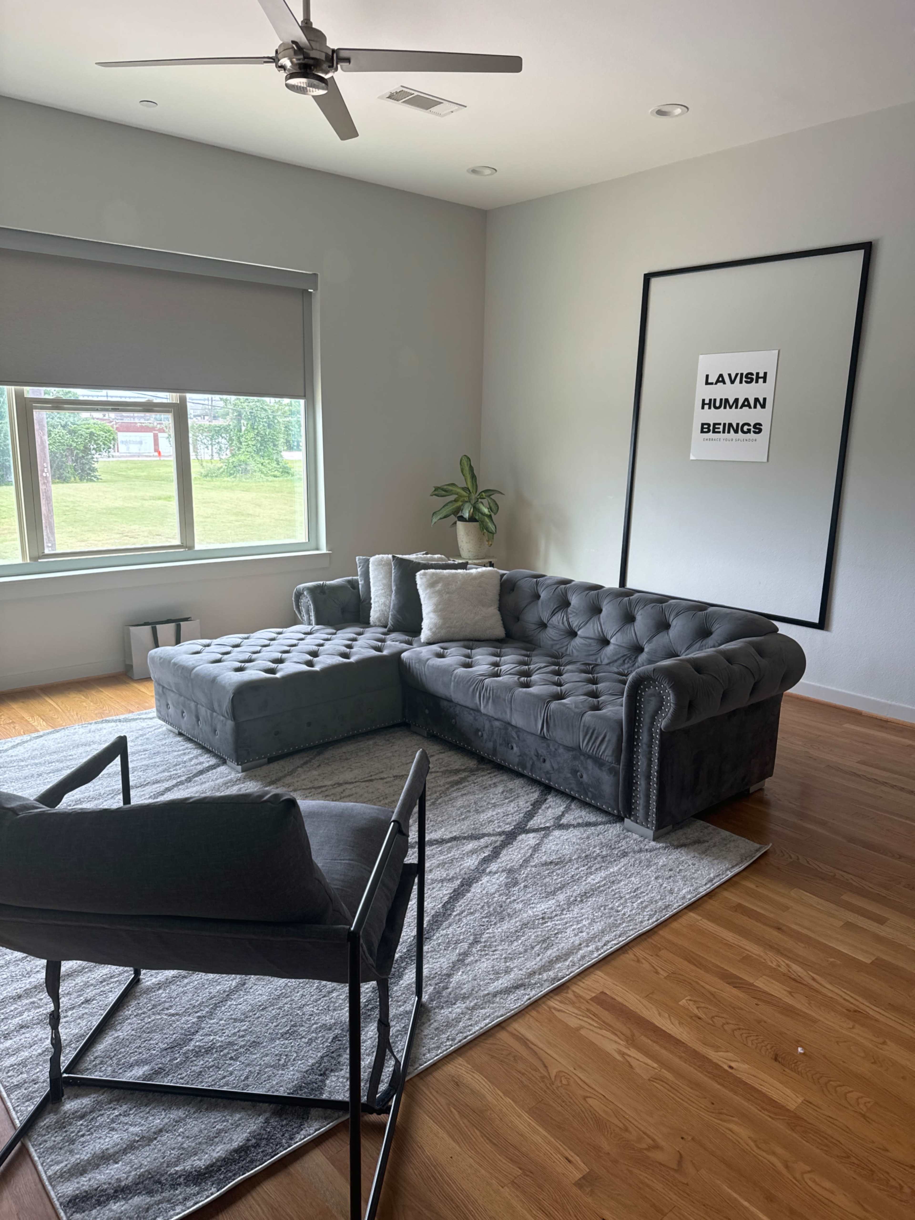 A modern living room featuring a gray tufted sectional sofa, a black metal chair, and a large window with a view of greenery.