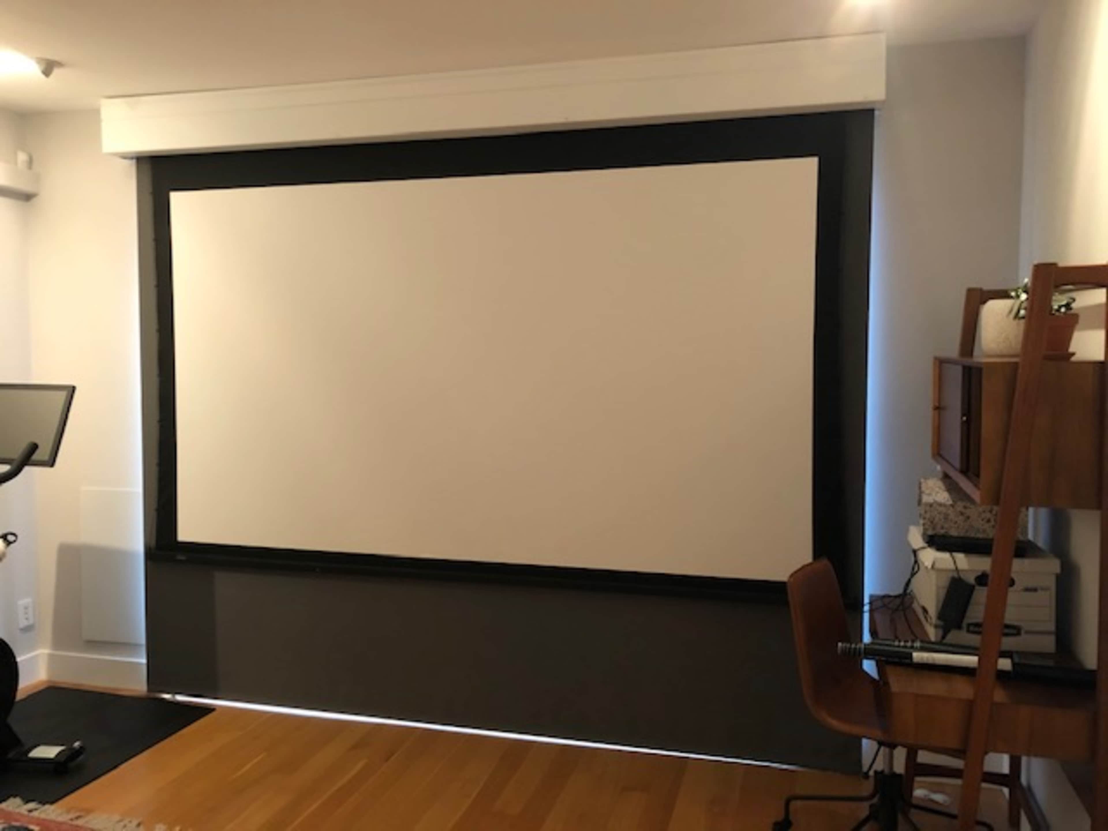 The image shows a room with a large retractable projection screen mounted on the wall, a wooden desk with various items, and a chair nearby.