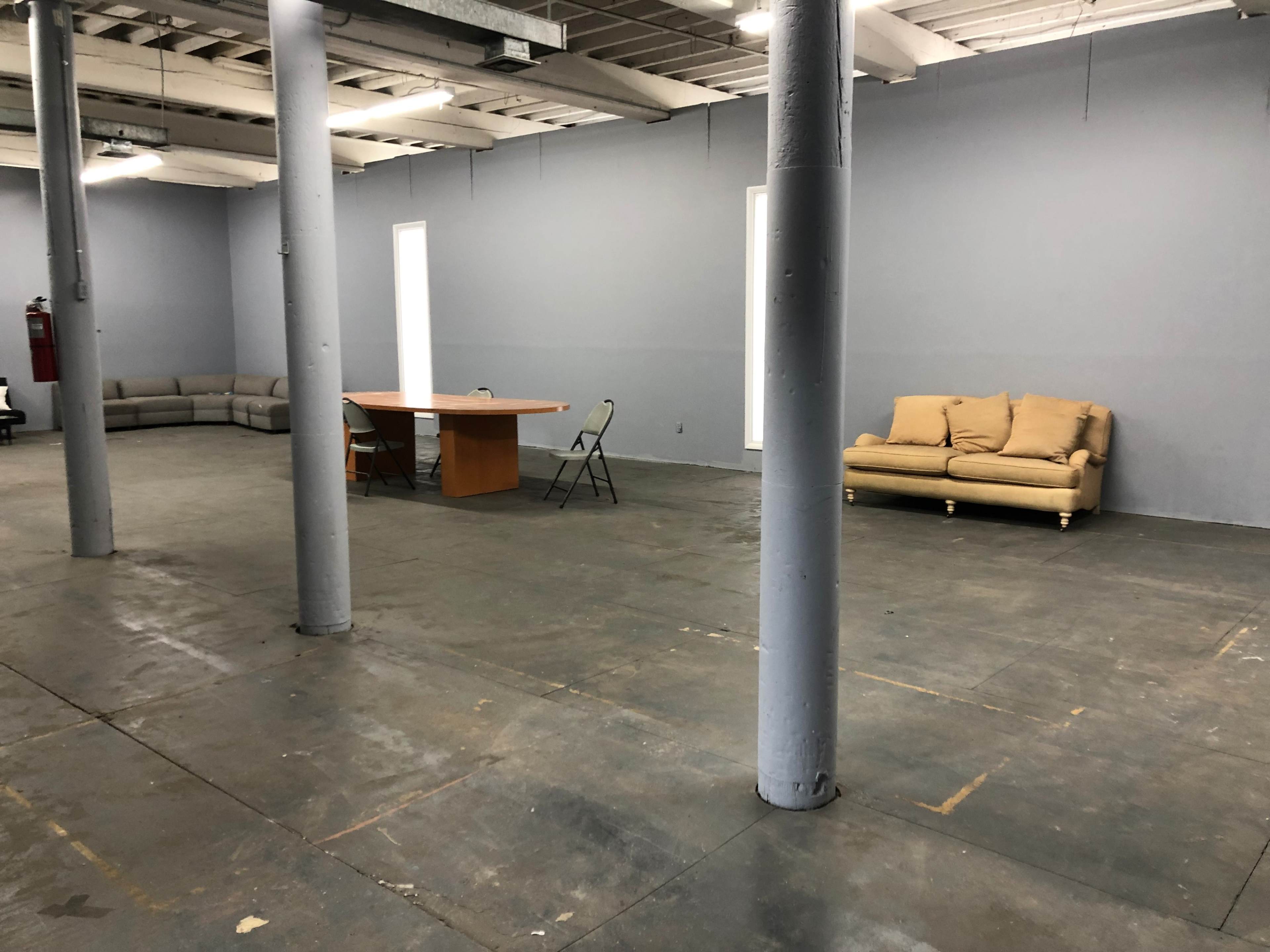 An empty industrial space with gray walls, two windows, a table with chairs, and a couch.