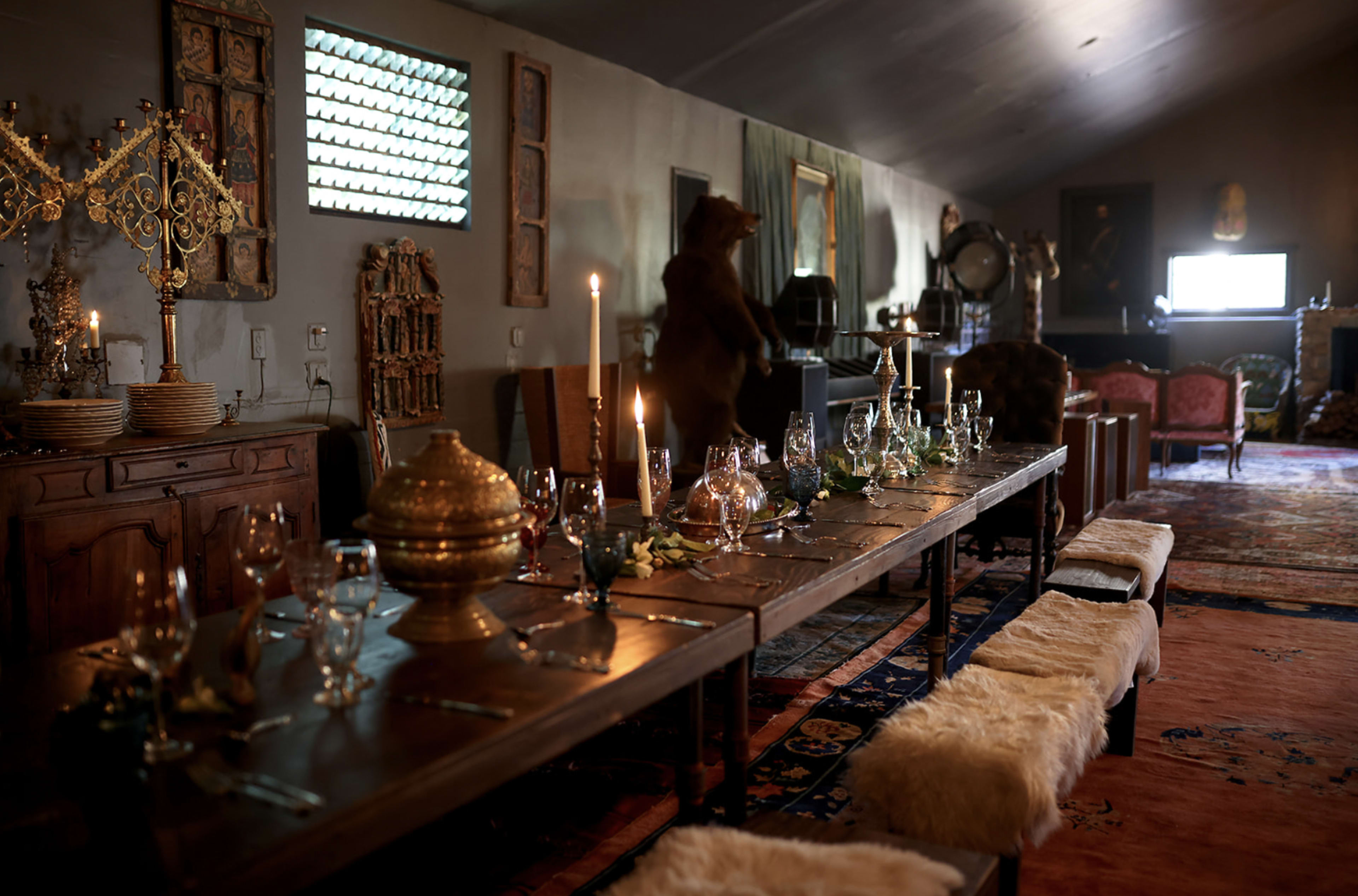 A long dining table set with glassware and candle holders is positioned in a rustic, dimly lit room adorned with various decorative items and furniture.