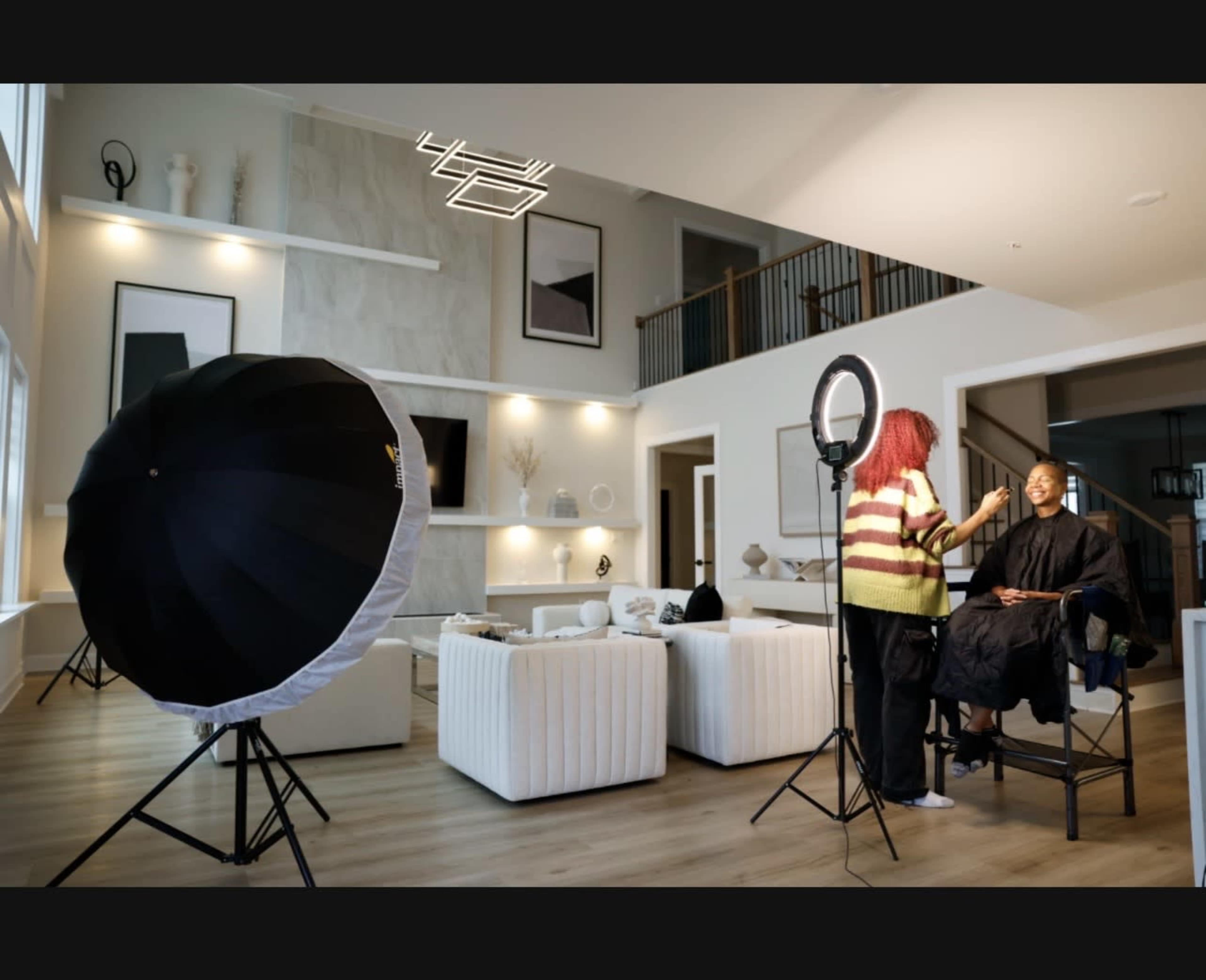 A makeup artist applies makeup to a seated client in a modern, well-lit living room.