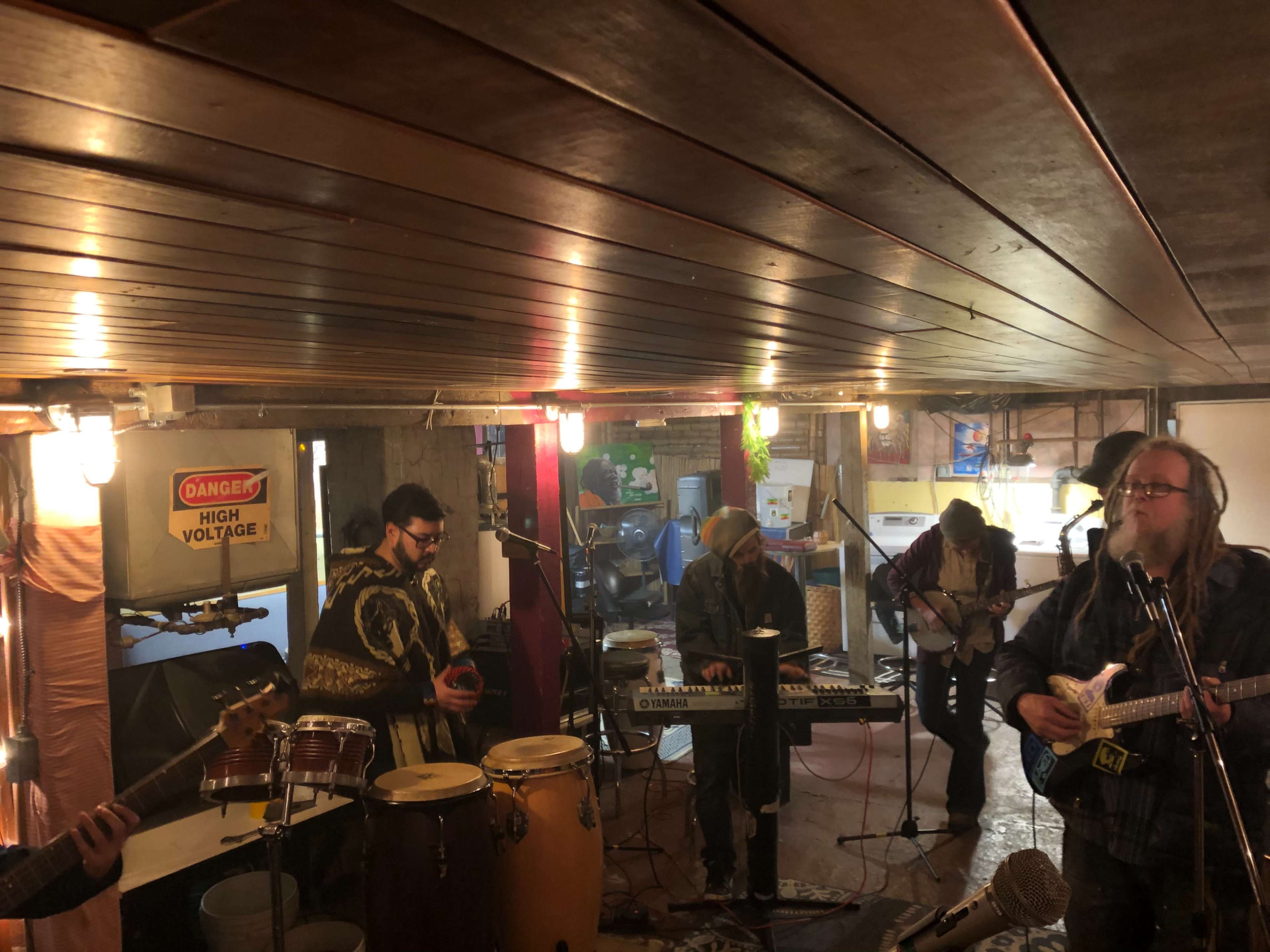 A group of musicians plays instruments in a basement setting with warm lighting and exposed wooden beams.