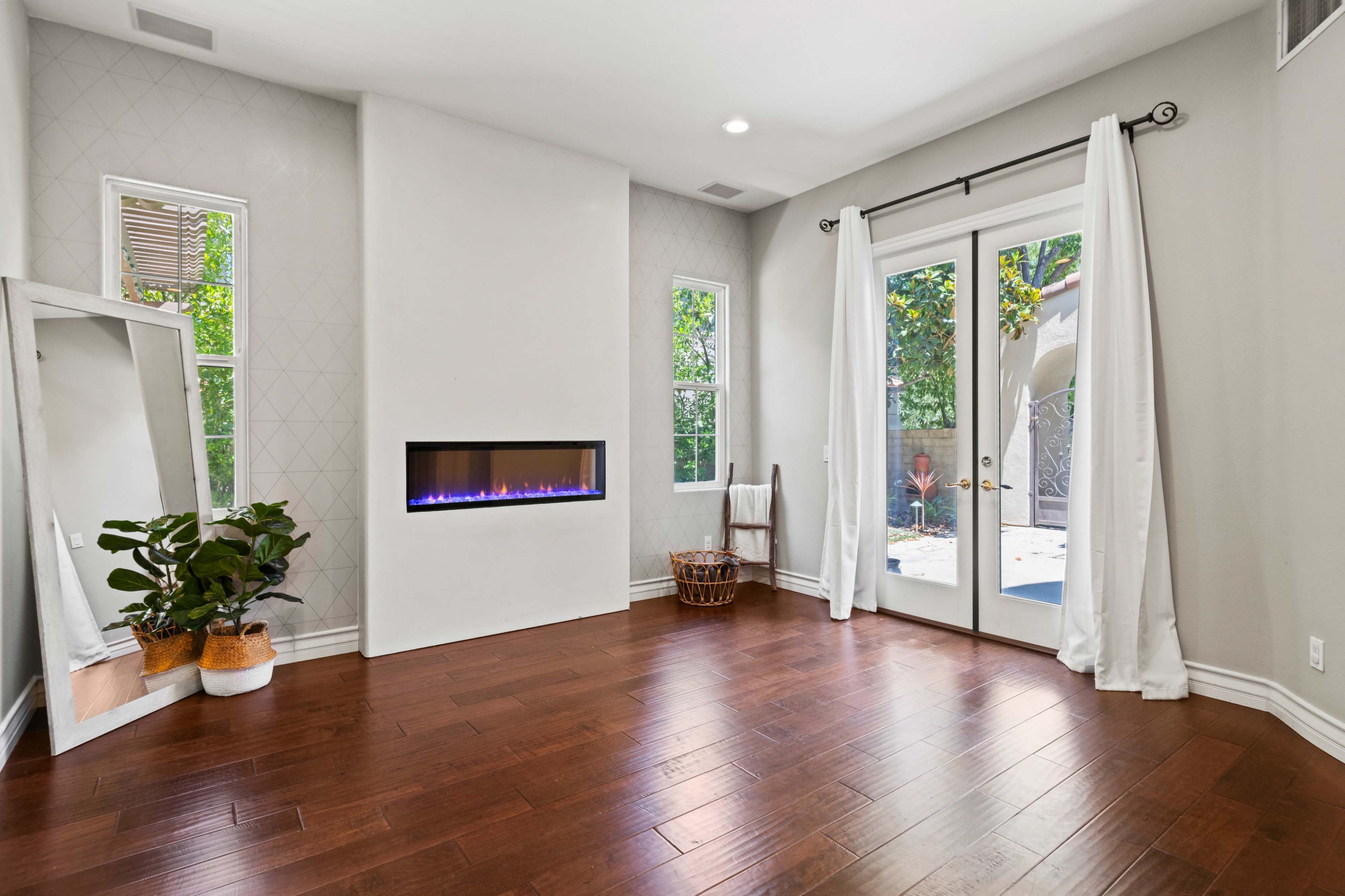 A room with hardwood floors, a modern electric fireplace, large windows, and a pair of doors leading outside, framed by white curtains.