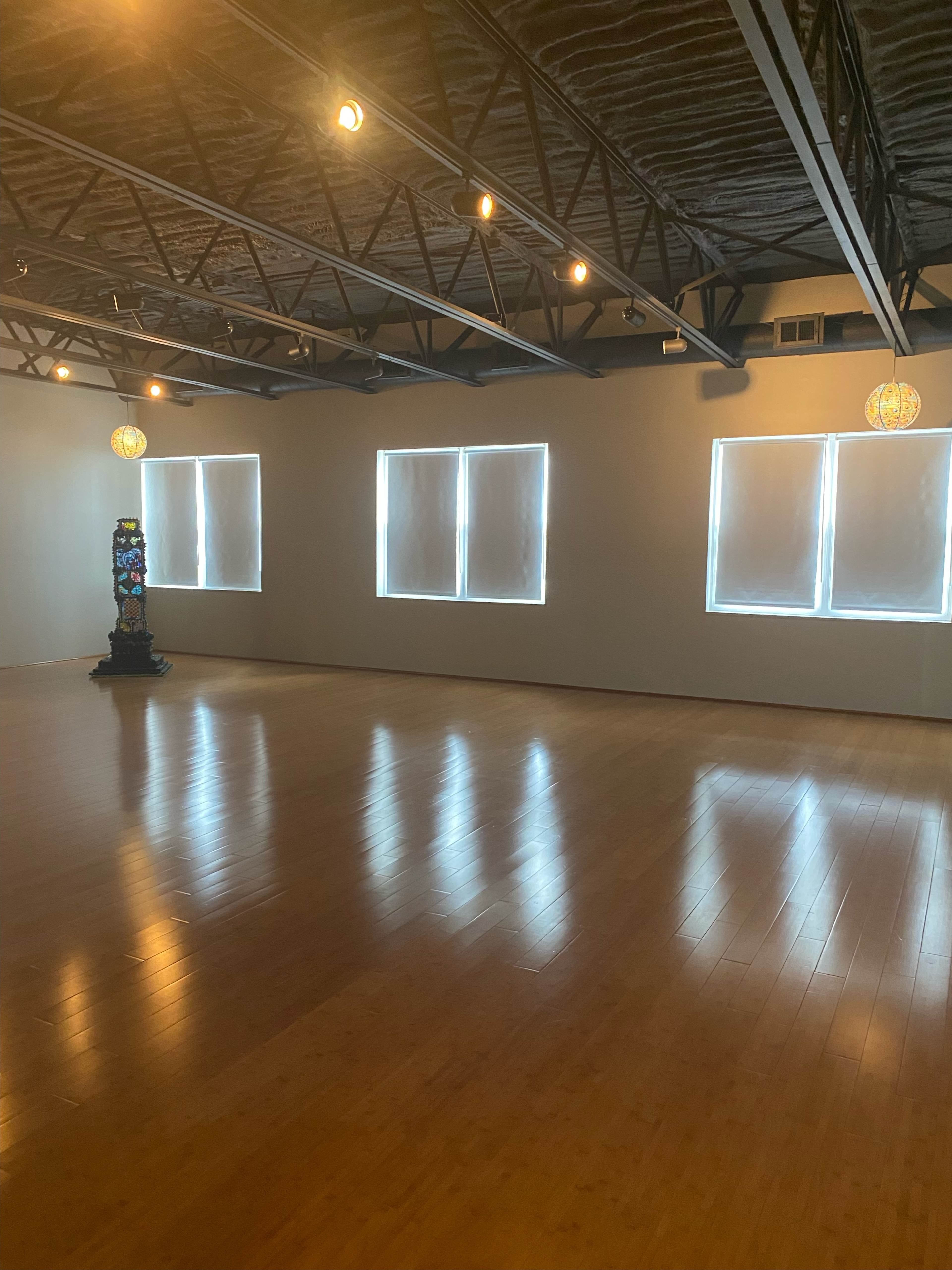 A spacious, empty room with wooden floors, large windows covered in white shades, and decorative hanging lights.