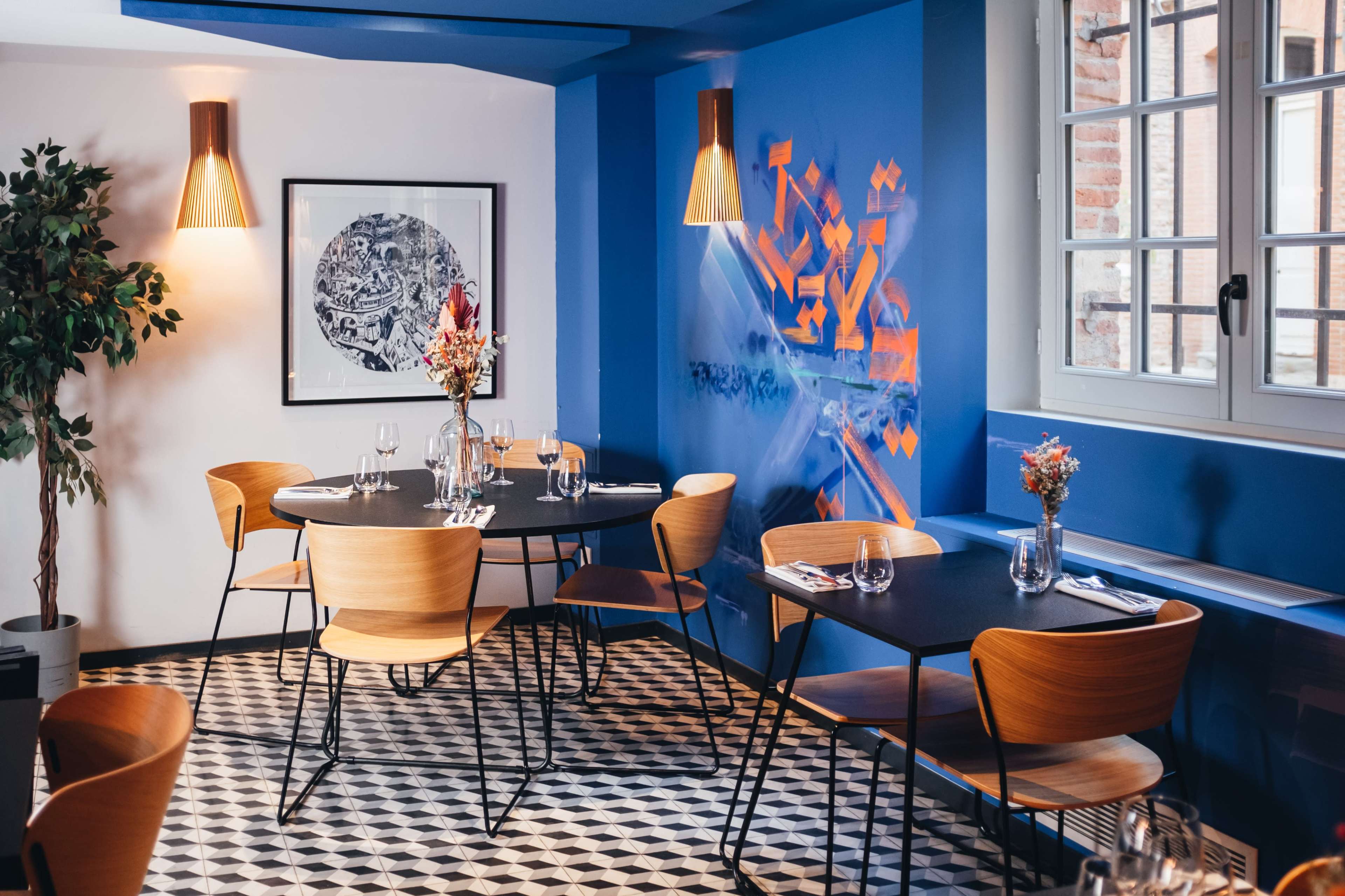 The image shows a modern restaurant interior featuring a circular table surrounded by wooden chairs, with blue walls adorned with artwork and large windows letting in natural light.