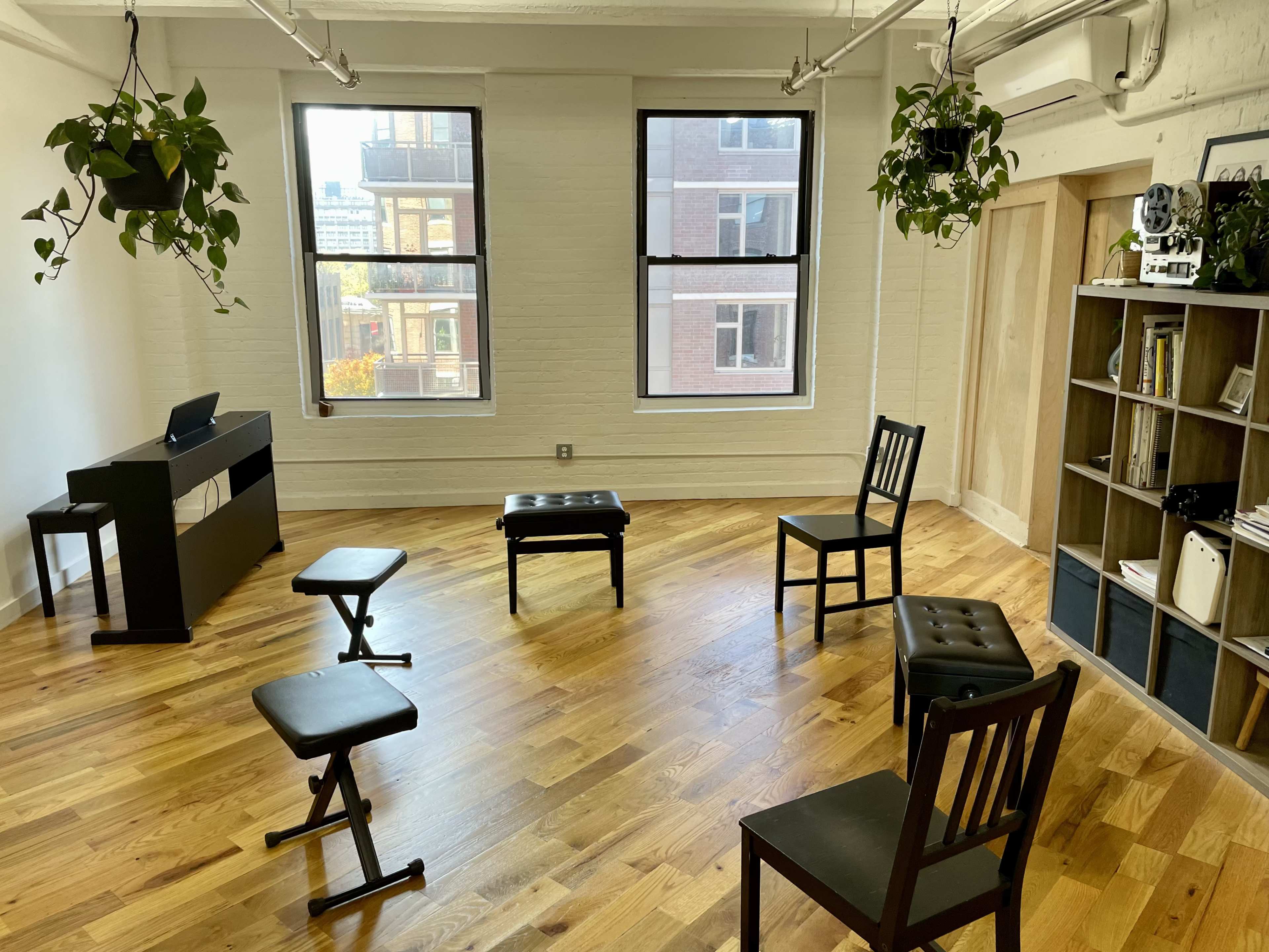 A sparsely furnished room with wooden floors, featuring a piano, several chairs, and decorative hanging plants.