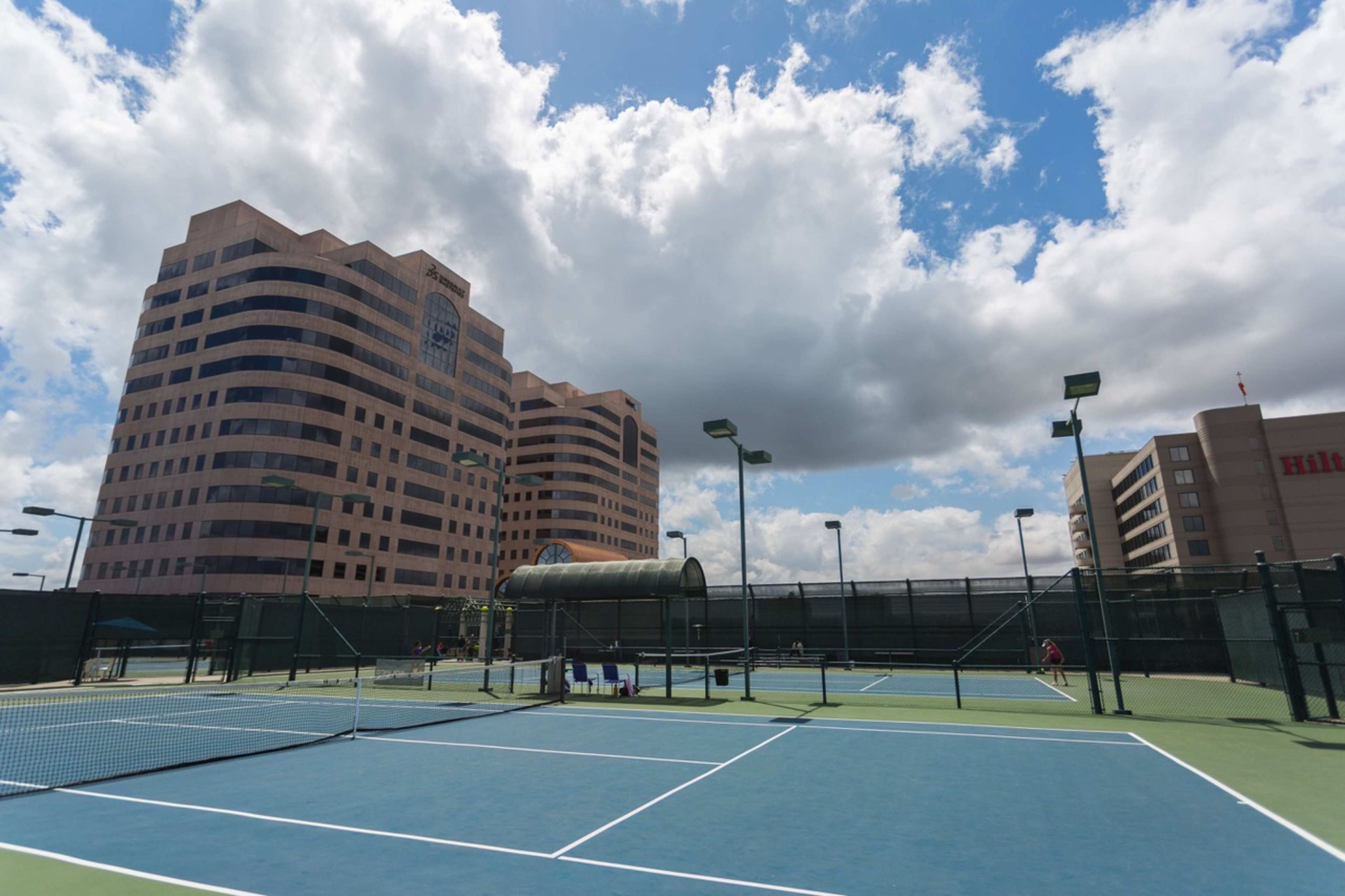 The image shows a tennis court with blue and green surfaces, surrounded by tall buildings under a partly cloudy sky.