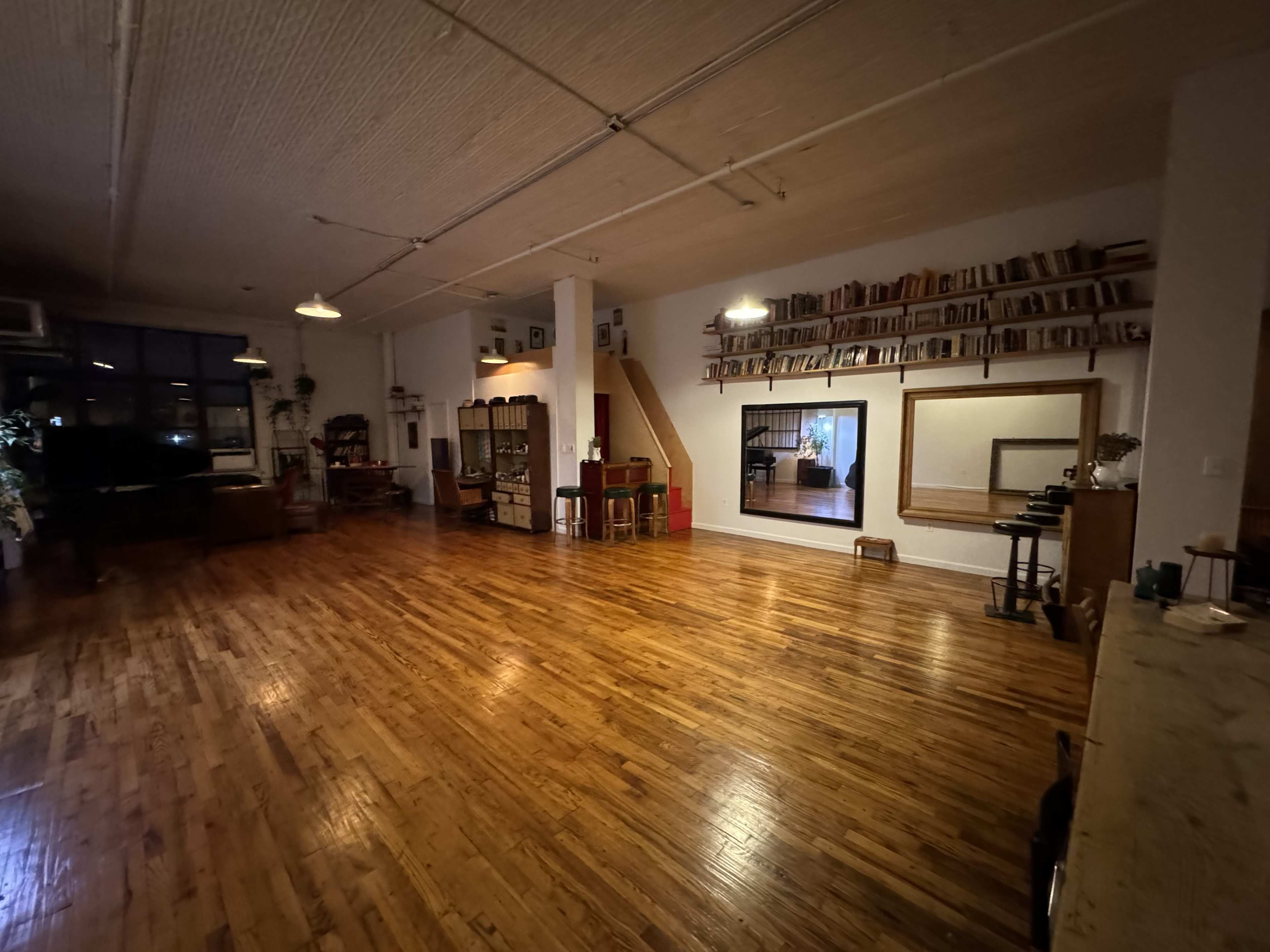 The image shows a large, empty room with wooden floors, shelves filled with books along one wall, and a mirror reflecting part of the space.