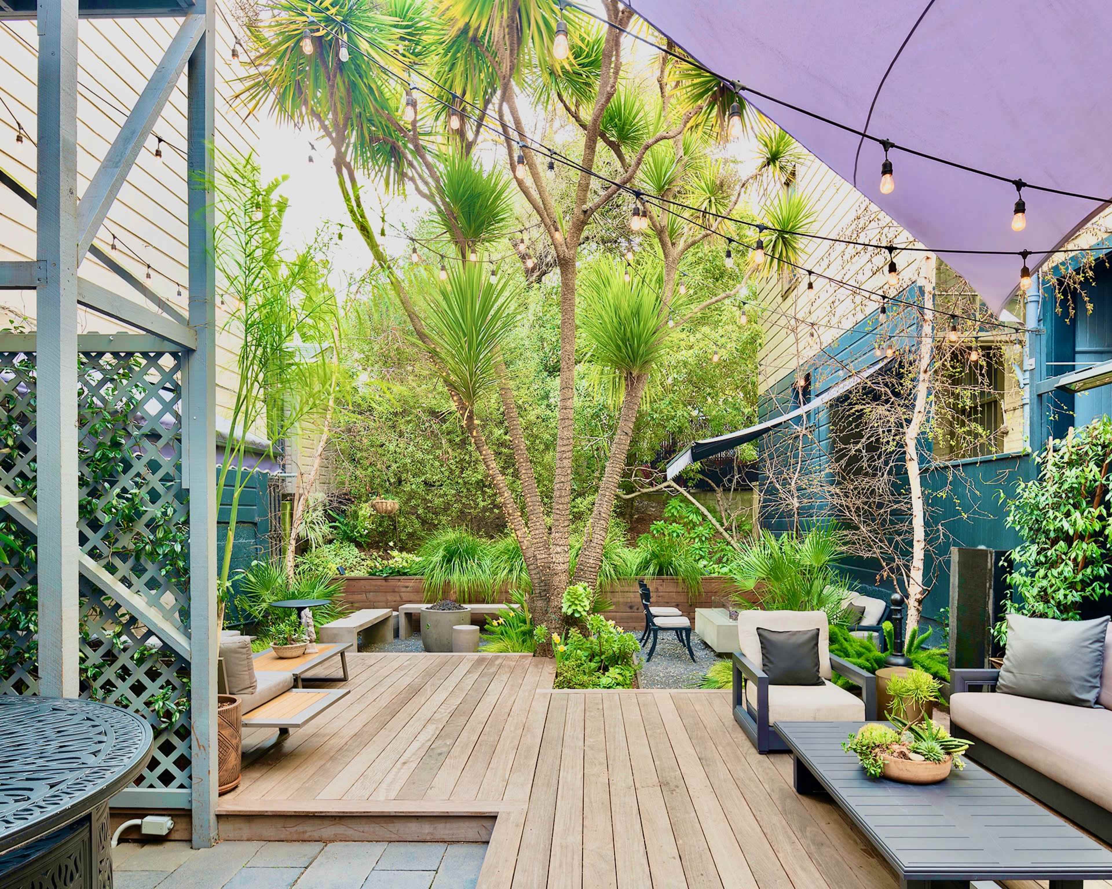 A wooden deck area is surrounded by lush greenery, featuring modern seating and decorative plants under purple awnings and illuminated with string lights.