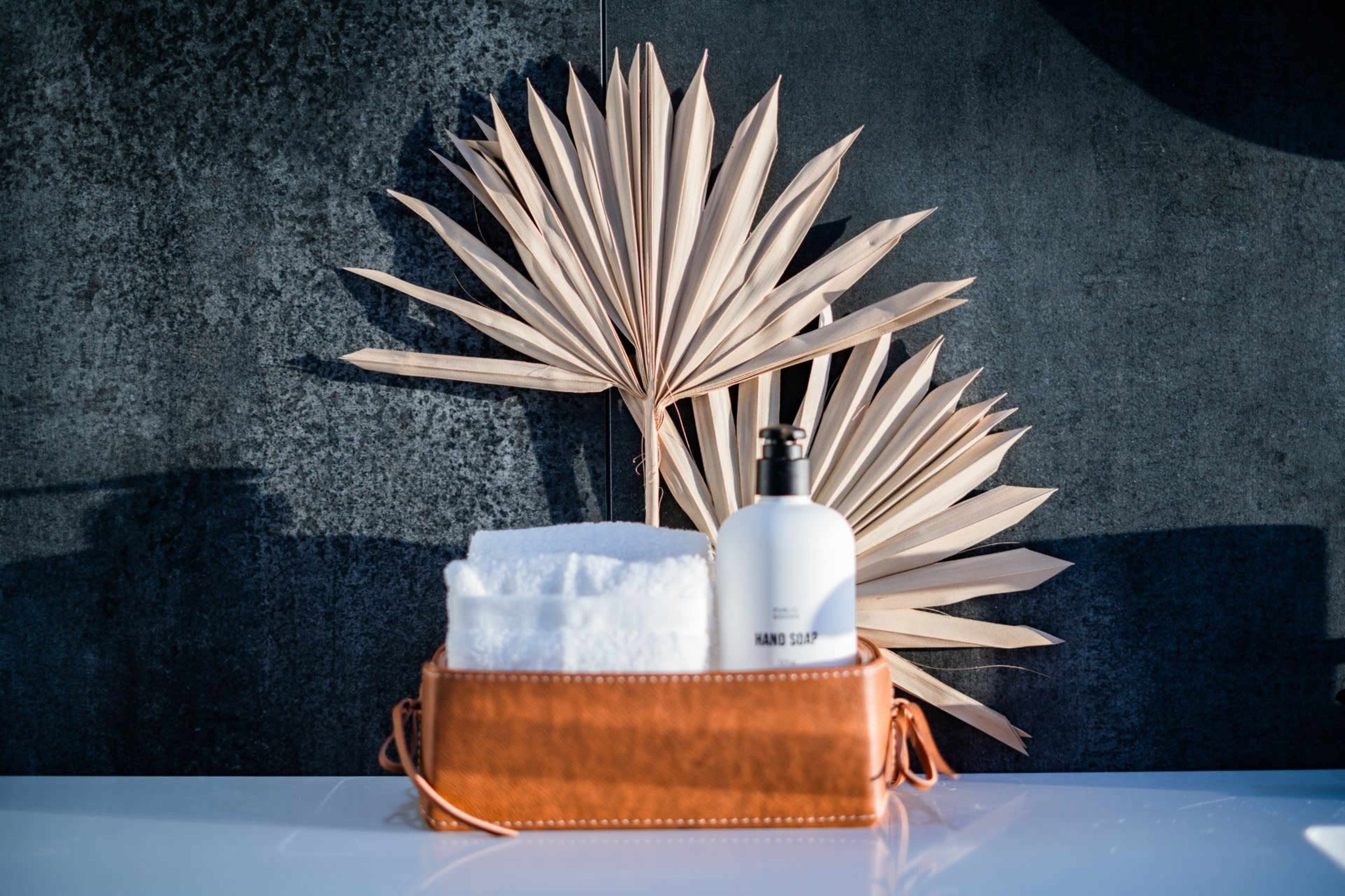 A basket containing a stack of white towels and a bottle of hand soap is placed against a backdrop of a palm leaf and a dark wall.