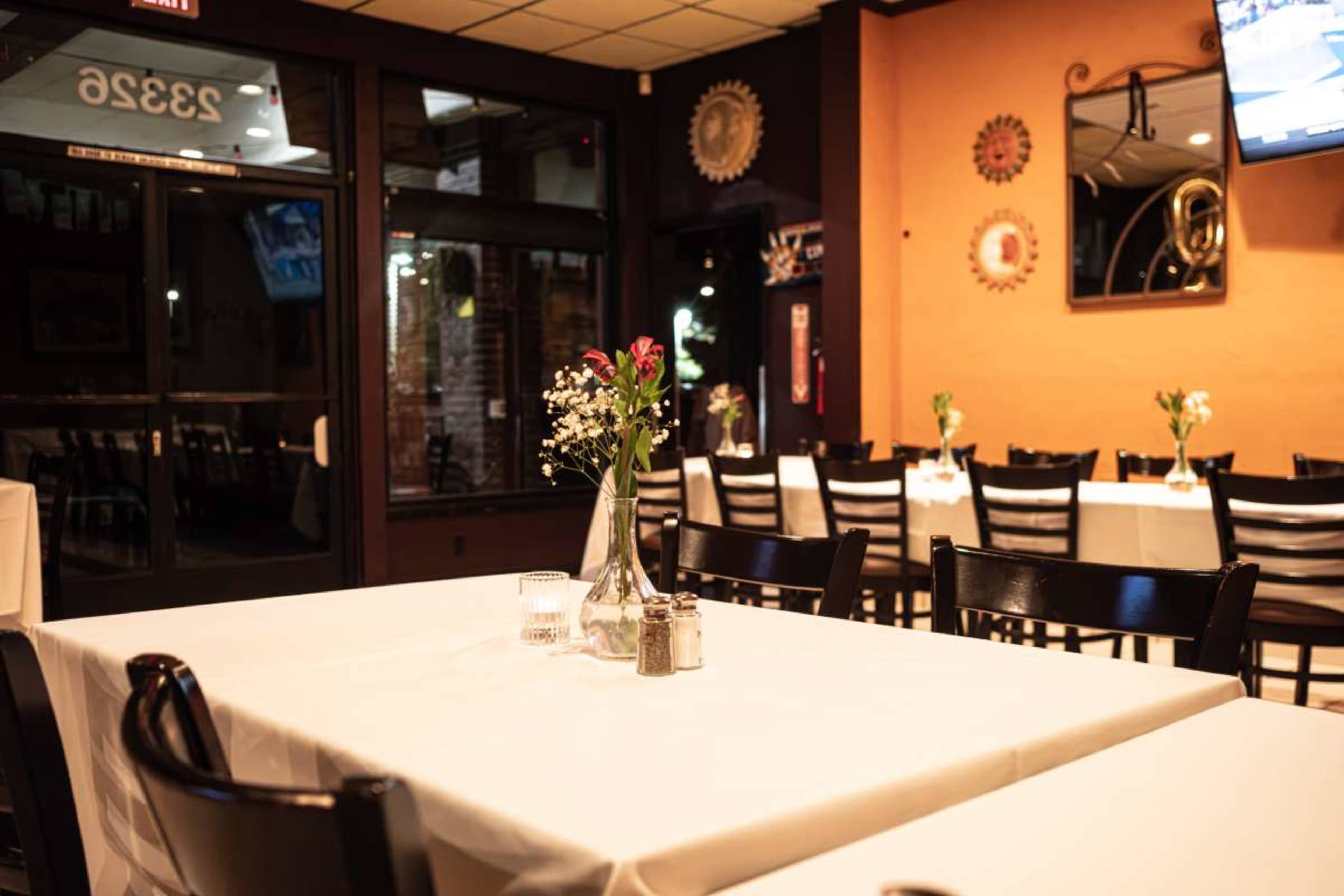 The image shows a semi-empty restaurant with tables set for dining, featuring white tablecloths and small flower arrangements in each vase.