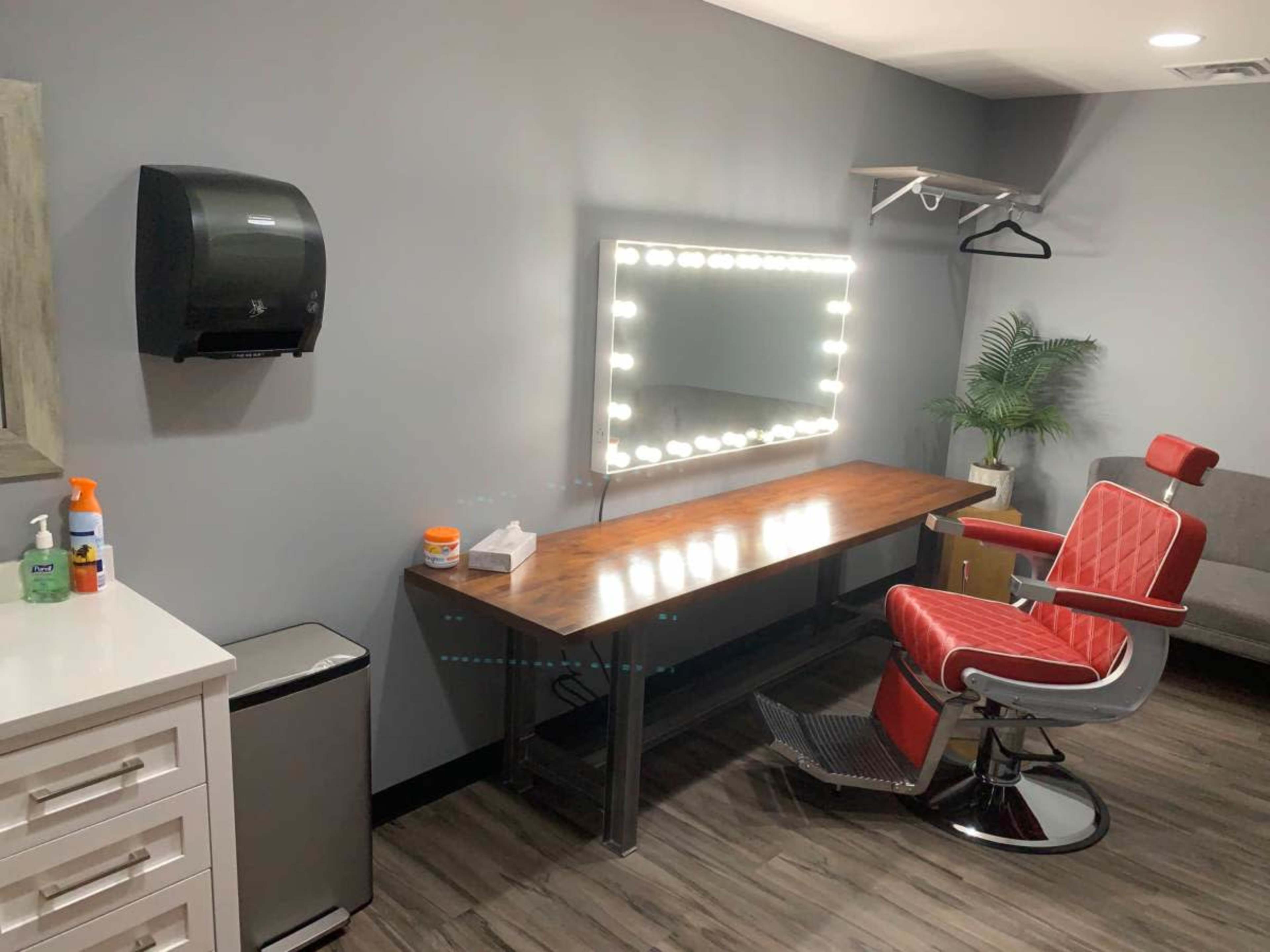 The image shows a modern barber or styling station featuring a red barber chair, a well-lit mirror surrounded by lights, a wooden countertop, and a small fridge in a neatly organized room.