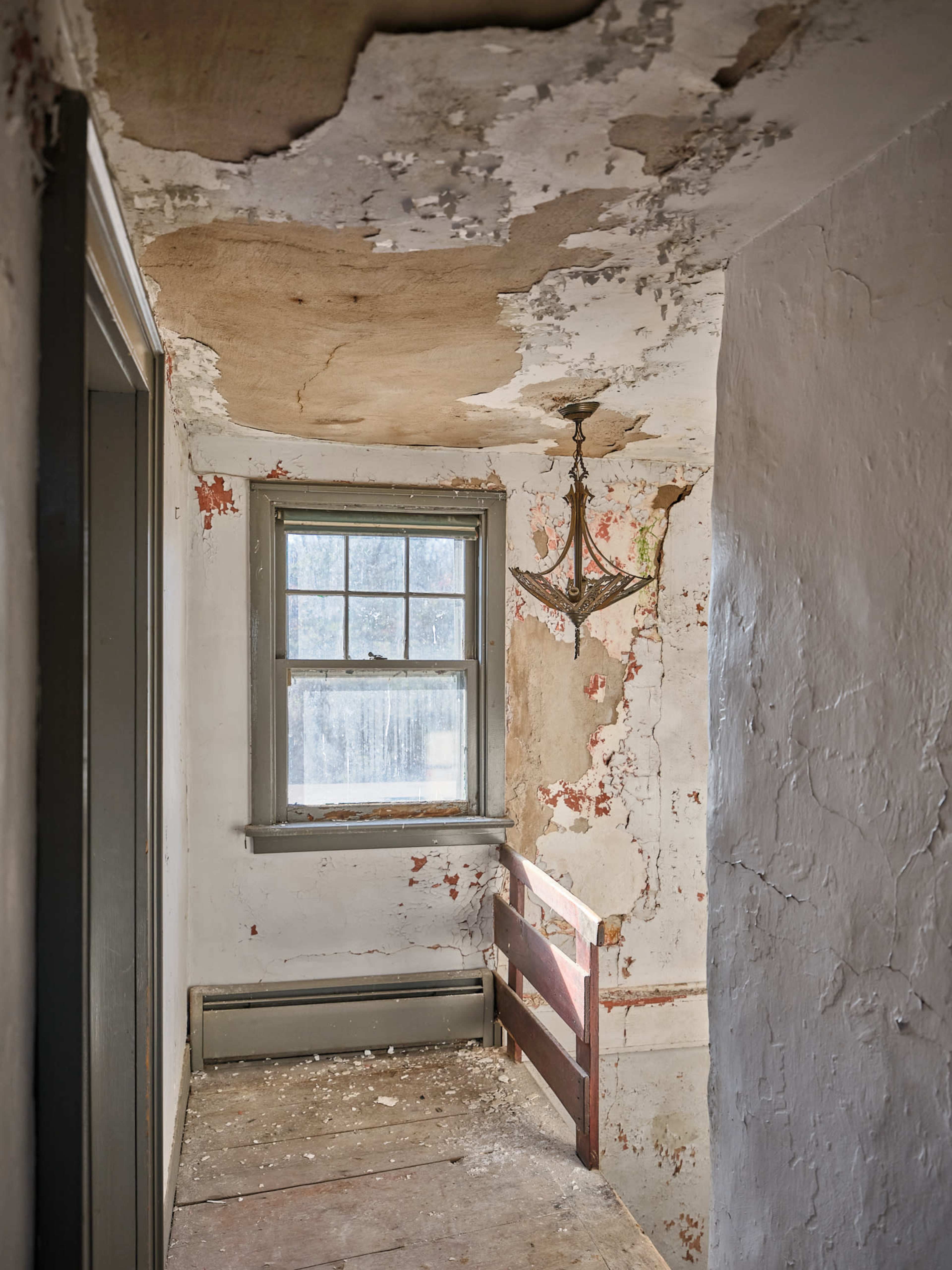 A dilapidated hallway with peeling paint, an old window, and a bare light fixture hanging from the ceiling.