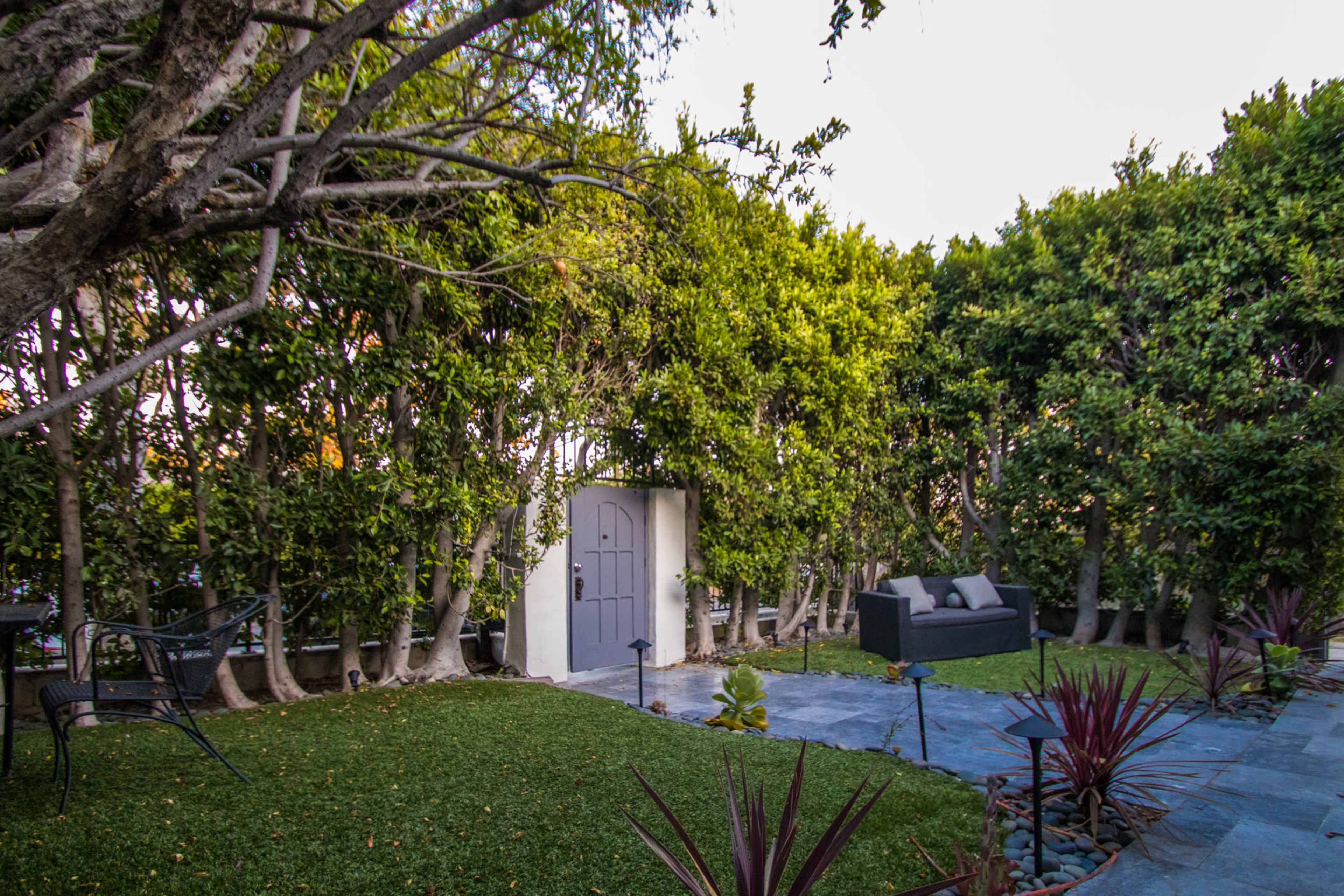 The image shows a landscaped garden featuring a small stone path, a neatly trimmed grassy area, a gray couch, and dense hedges surrounding the space.