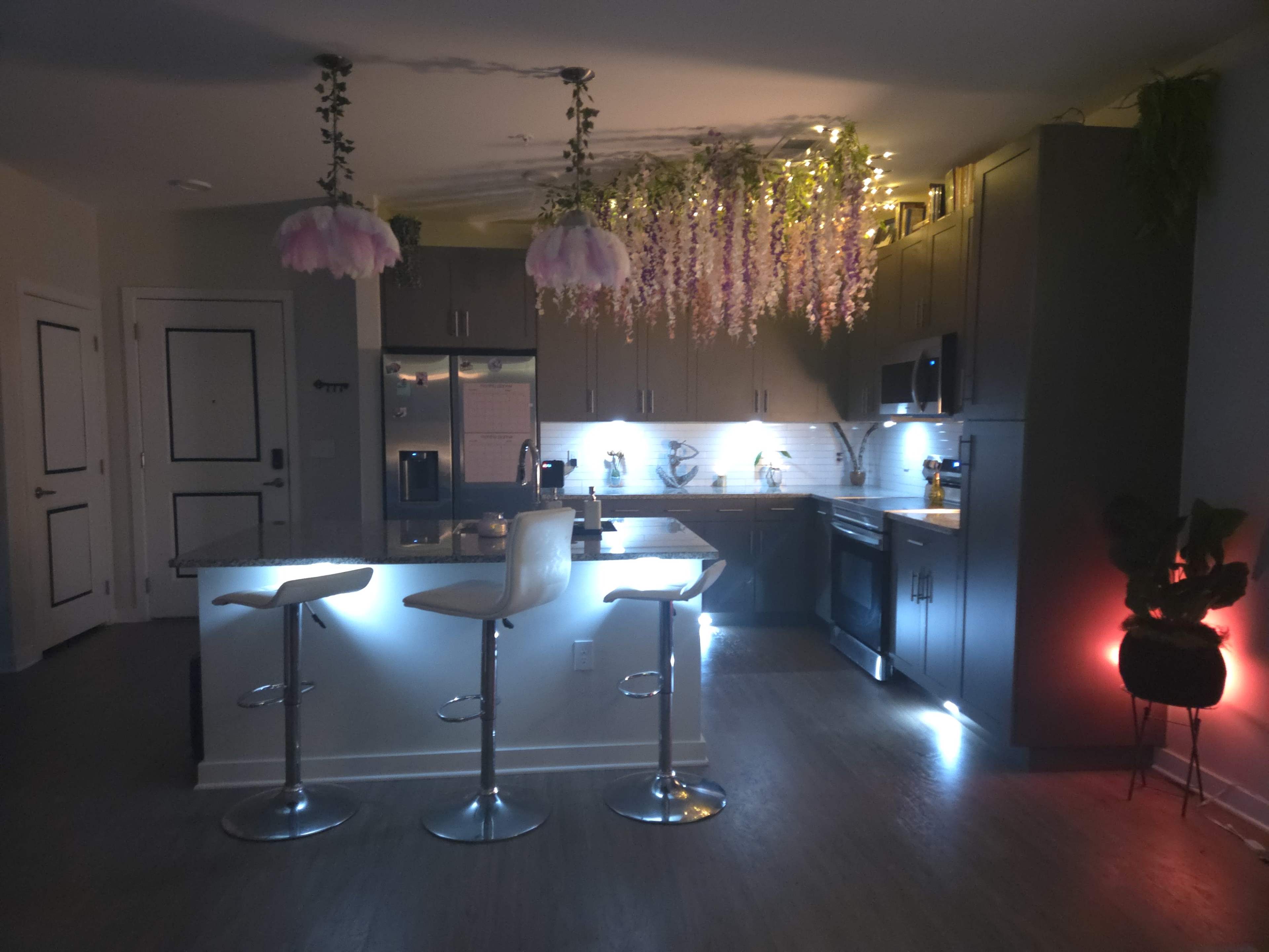The image shows a modern kitchen with pendant lights, bar stools, and decorative plants overhead, illuminated by soft lighting.