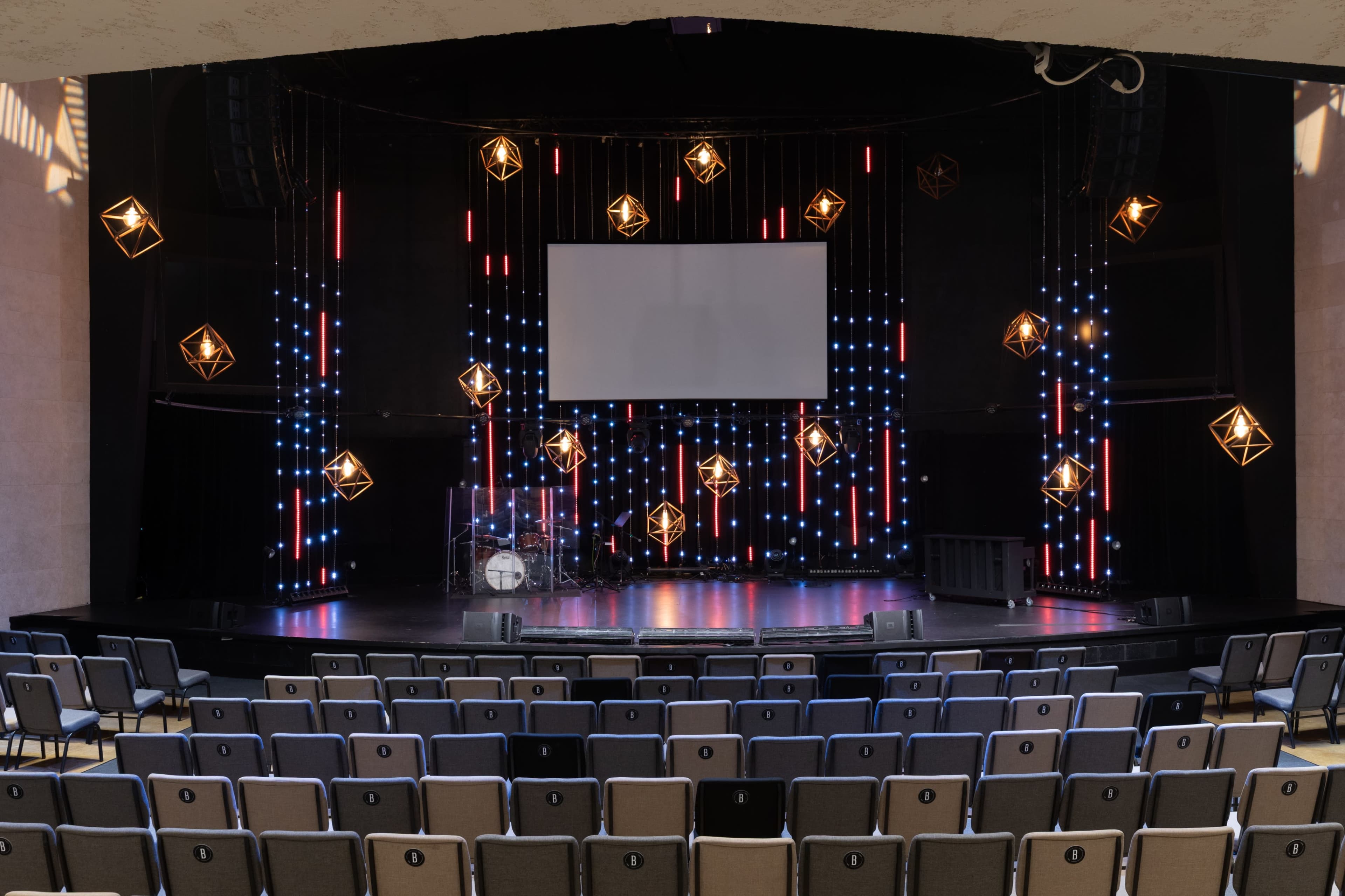 The image shows an empty theater stage with a large screen and hanging decorative lights above a circular band setup.