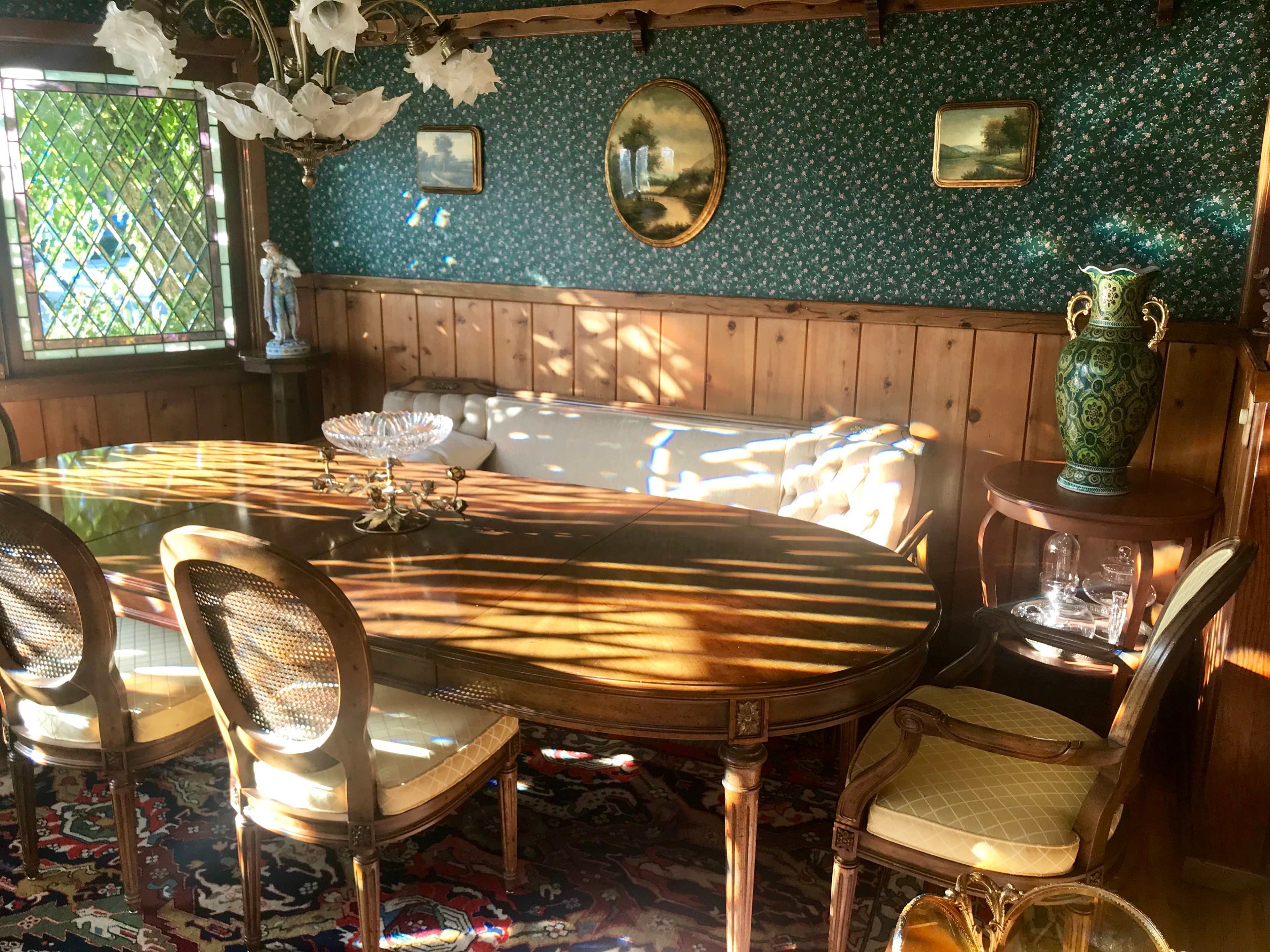 A wooden dining table surrounded by four chairs is positioned in a room with floral wallpaper, wooden paneling, and a vase on a side table.
