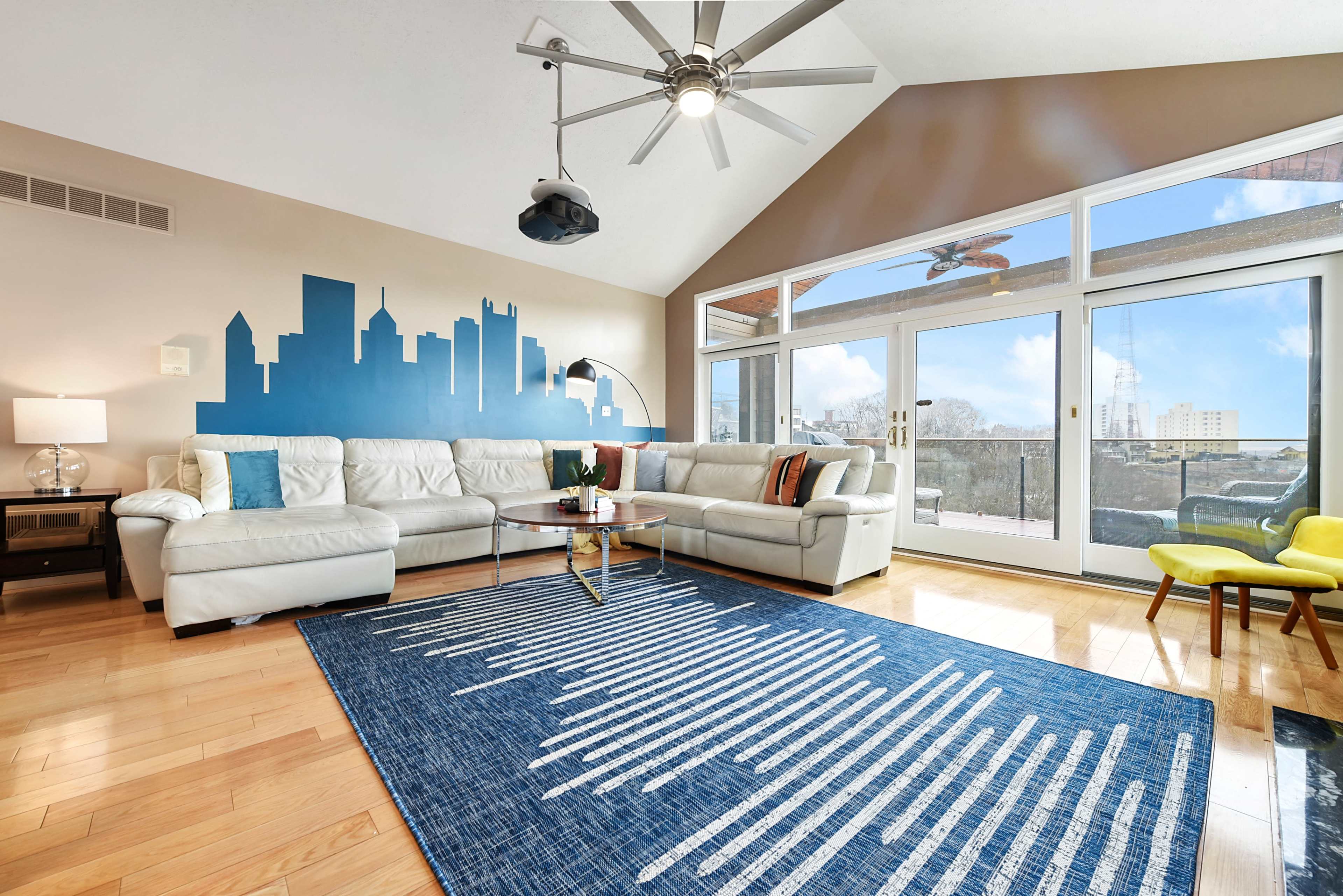 A modern living room features a large white sectional sofa, a blue skyline mural on the wall, and a stylish area rug on hardwood flooring.