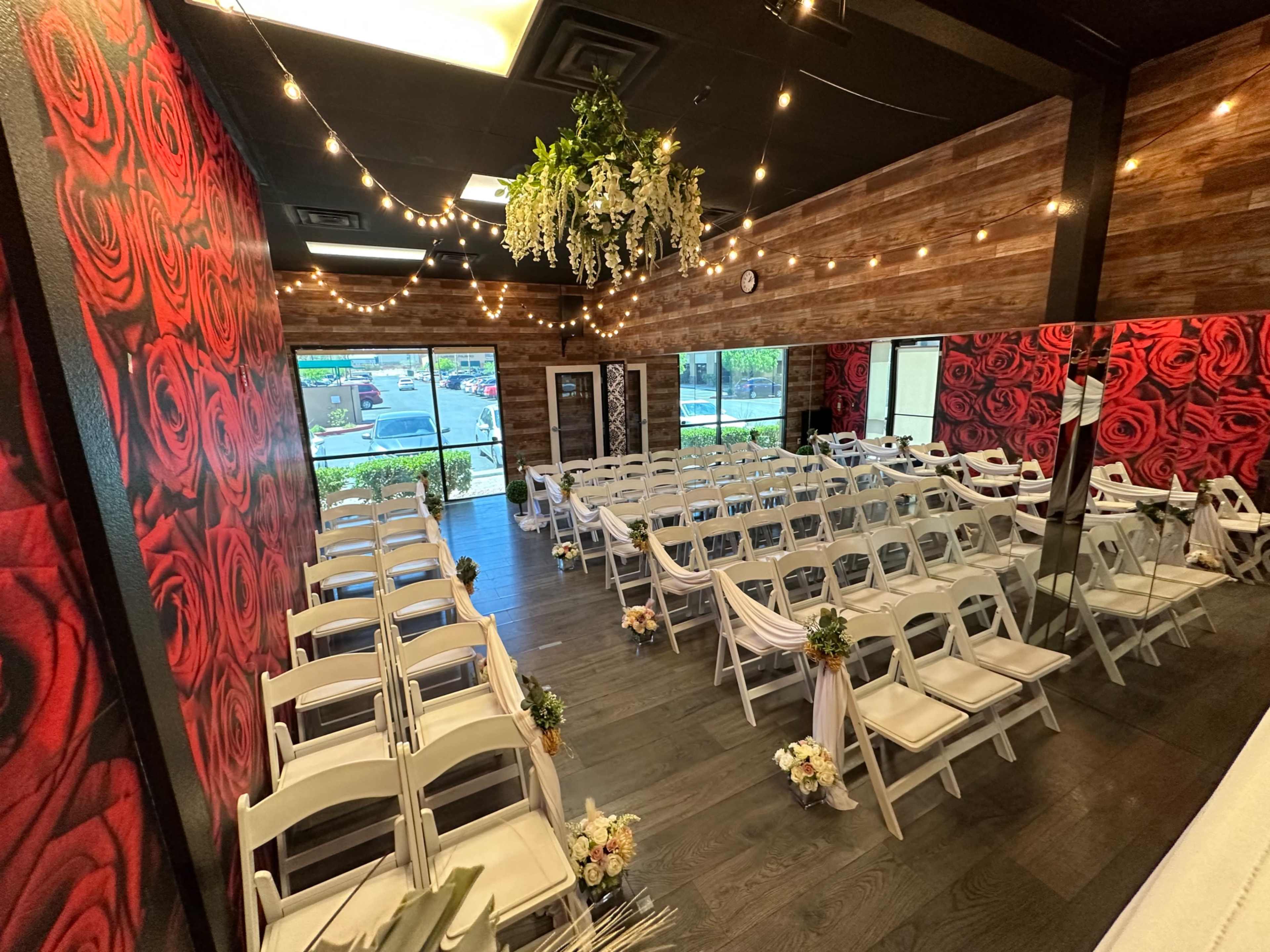A wedding venue features rows of white chairs arranged in front of a floral backdrop, with string lights overhead and floral decorations along the aisle.