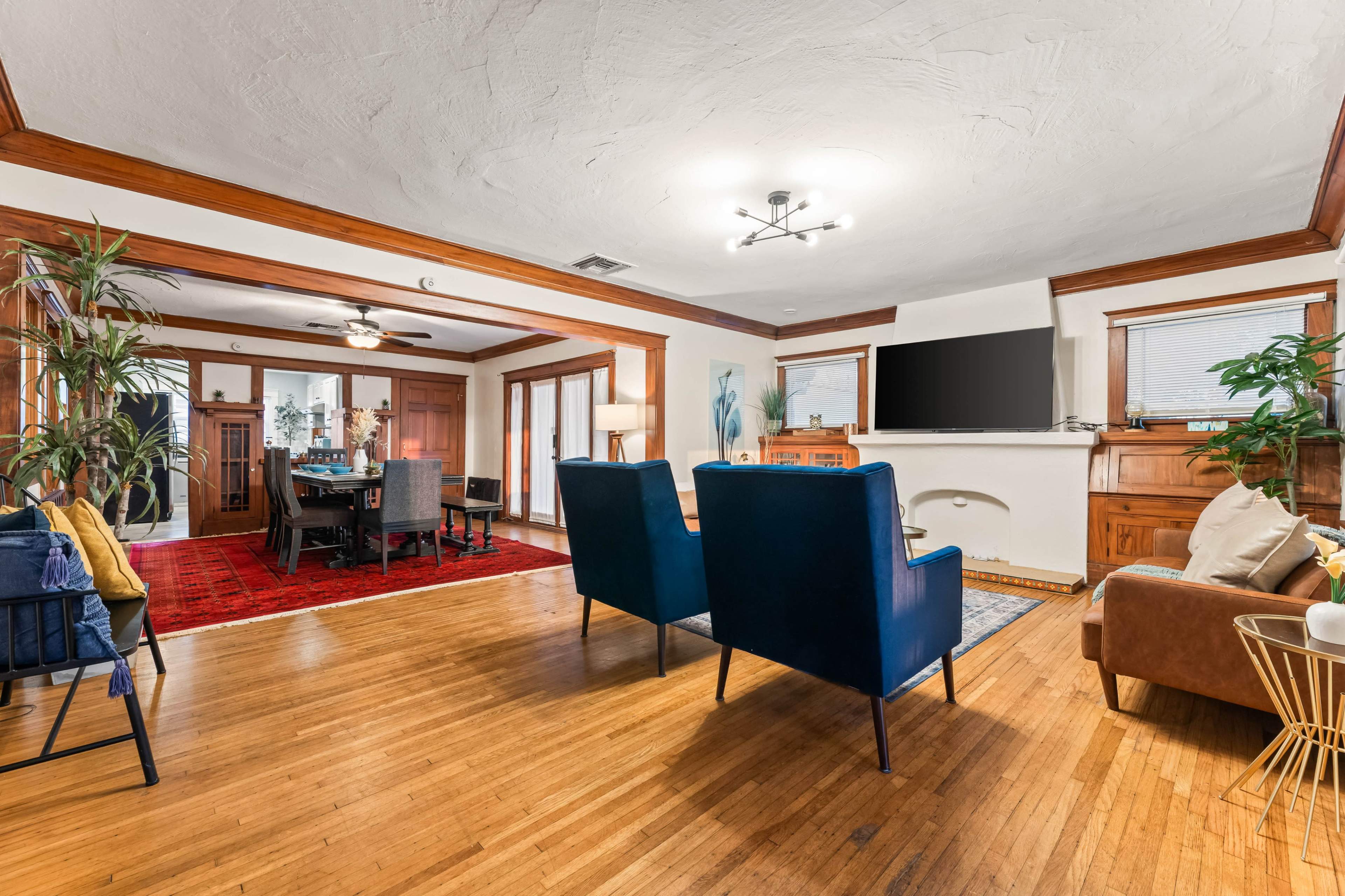 A spacious living room with hardwood floors features two blue armchairs facing a television, surrounded by plants and open to a dining area with a red rug.