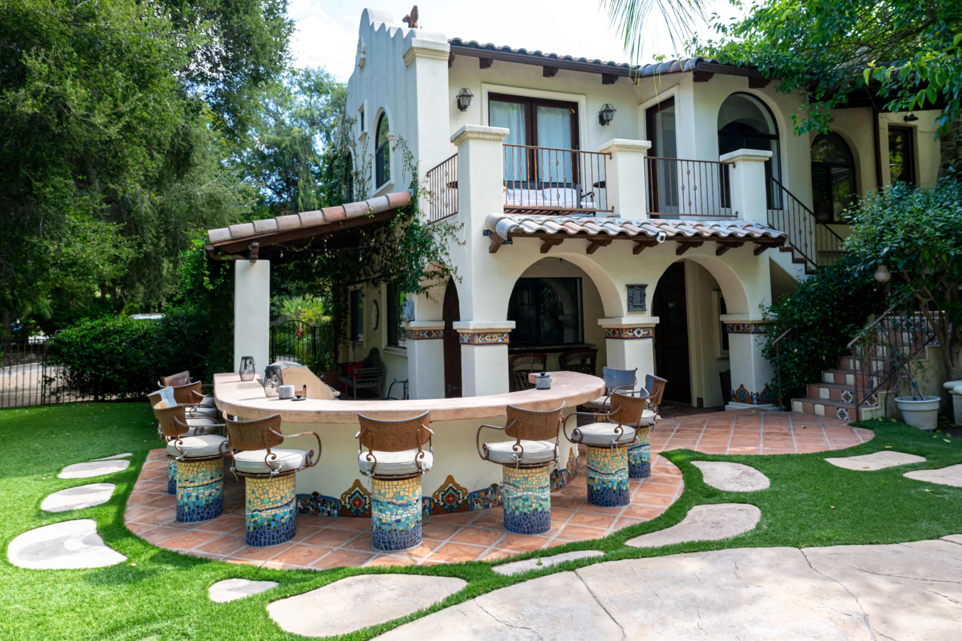 The image shows a colorful outdoor bar area with tiled seating, set against a two-story house surrounded by greenery.