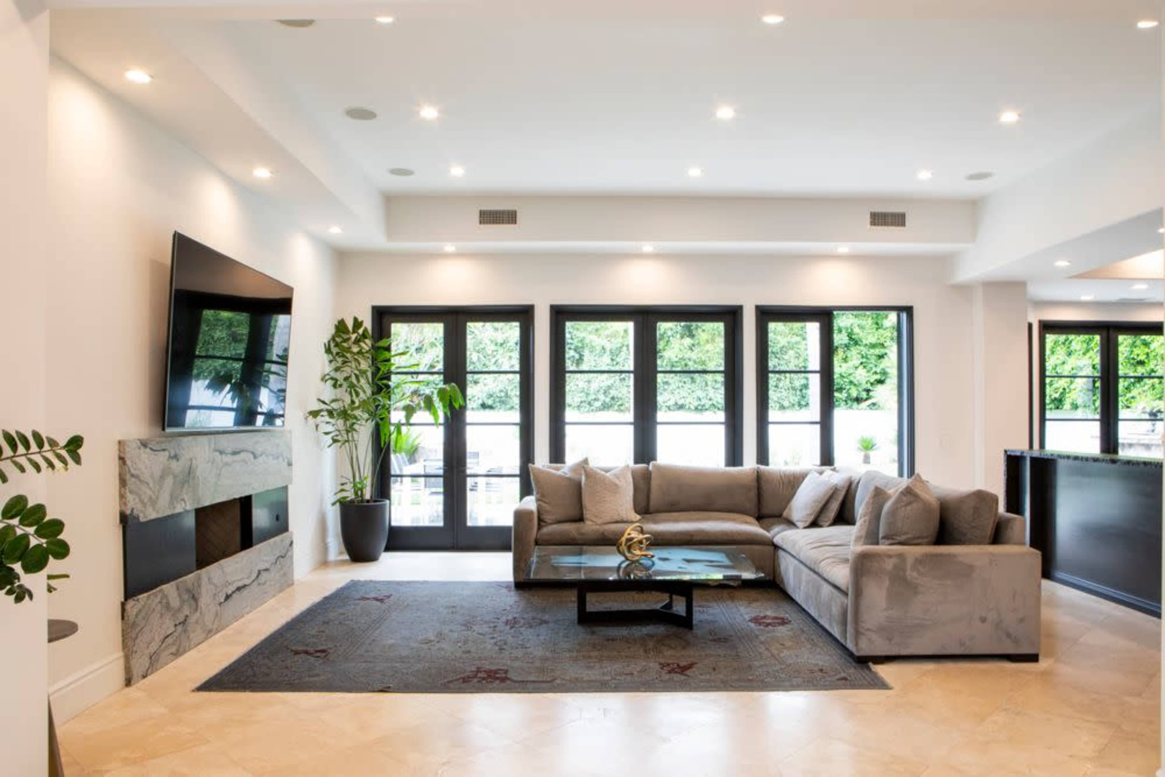 A modern living room with a large gray sectional sofa, a marble feature wall, a flat-screen TV, and large black-framed windows.