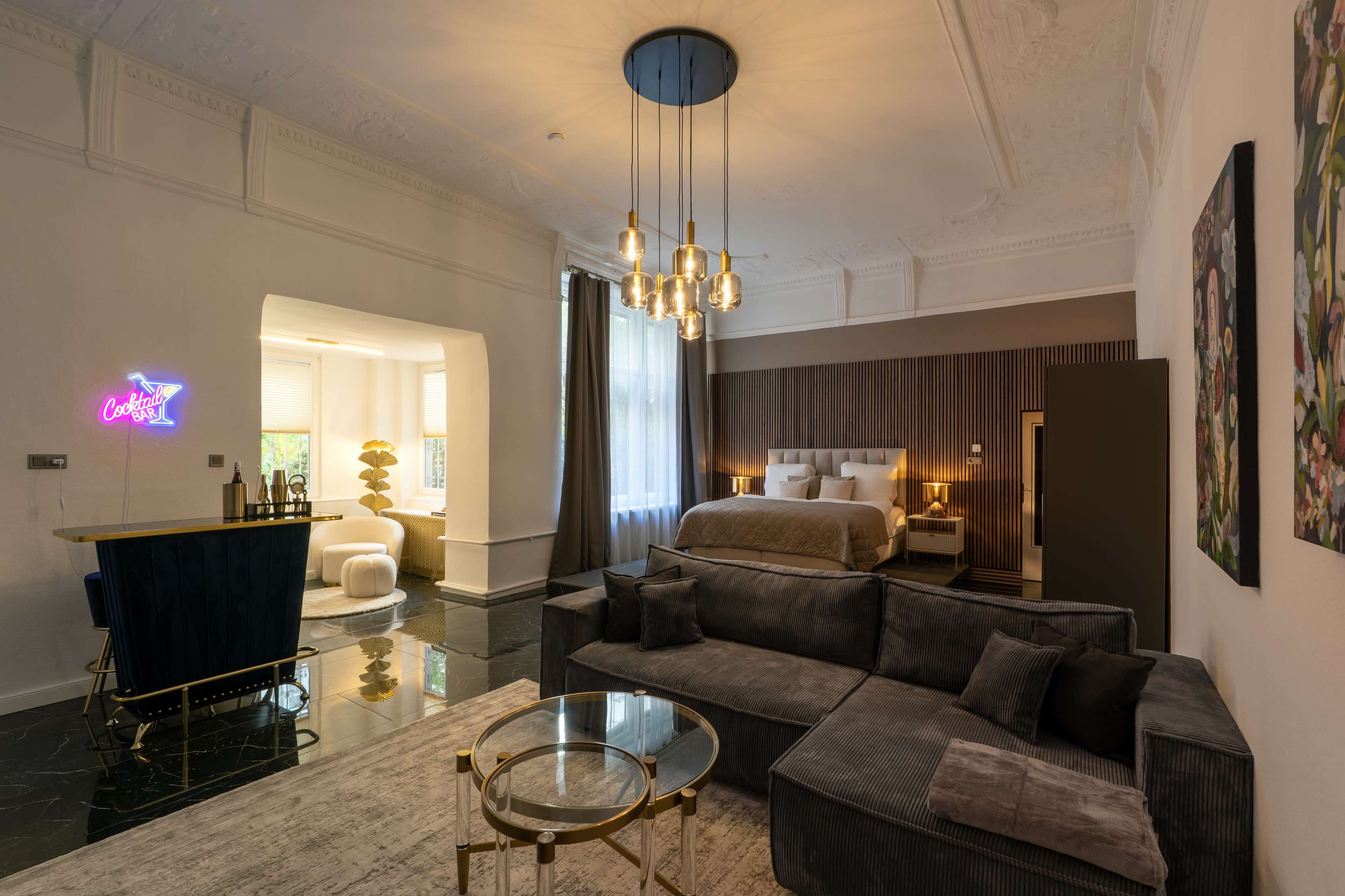 The image shows a stylish hotel room featuring a seating area with a gray sofa, a round coffee table, a bed with beige linens, and decorative wall art, illuminated by a modern chandelier.