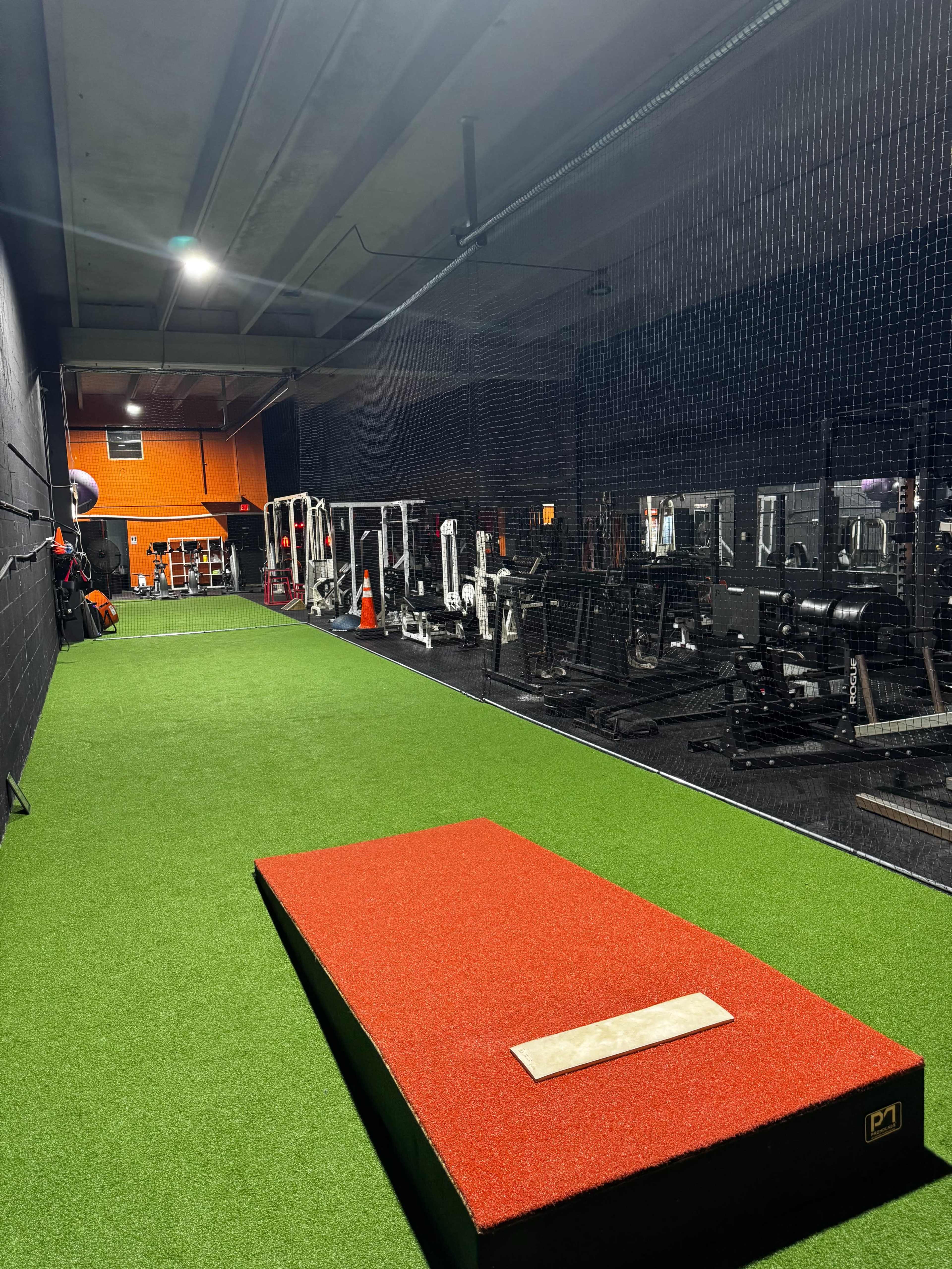 The image shows a gym featuring a bright green astroturf area with a red pitching mound, surrounded by various strength training equipment and mirrors.