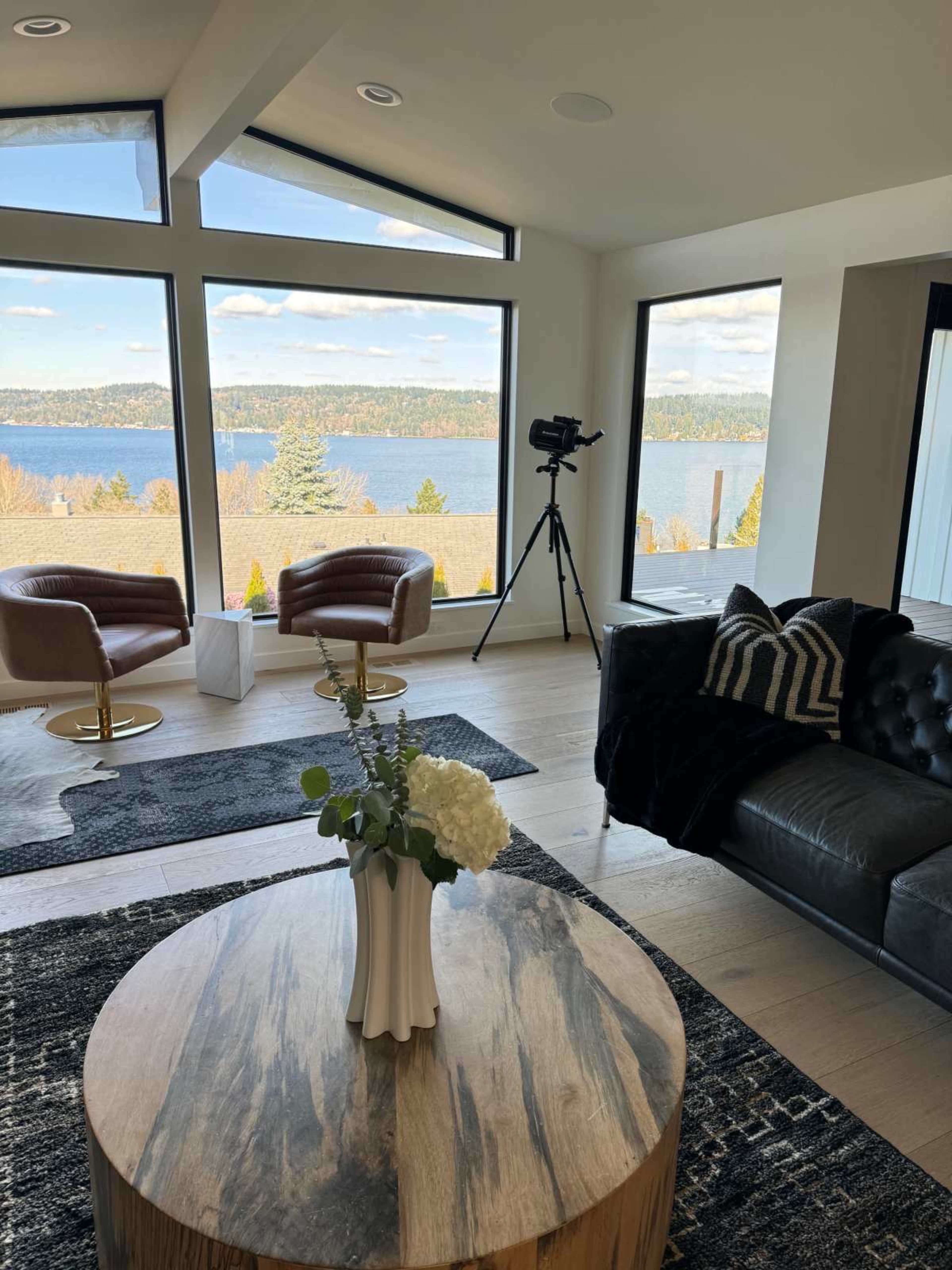 A modern living room features large windows overlooking a lake, with two armchairs, a telescope, and a wooden coffee table at the center.