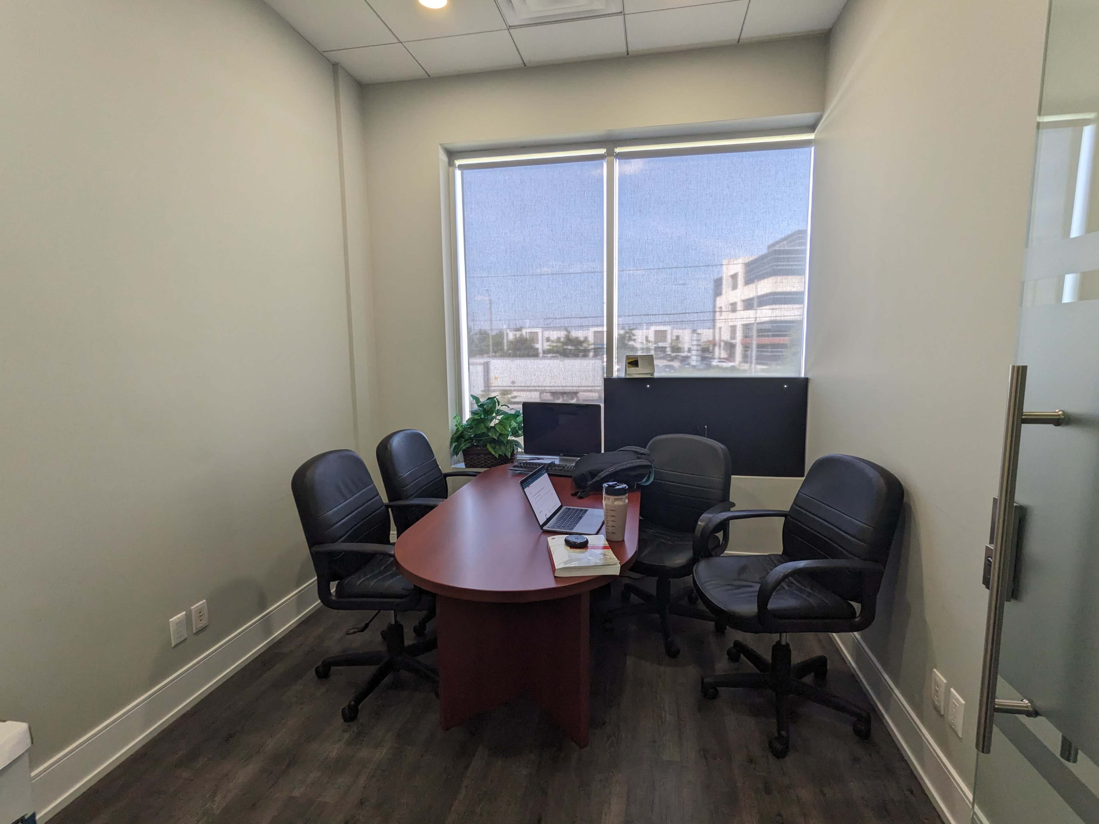 A small office with a round table and four black chairs, featuring a laptop and a potted plant, and large windows letting in natural light.